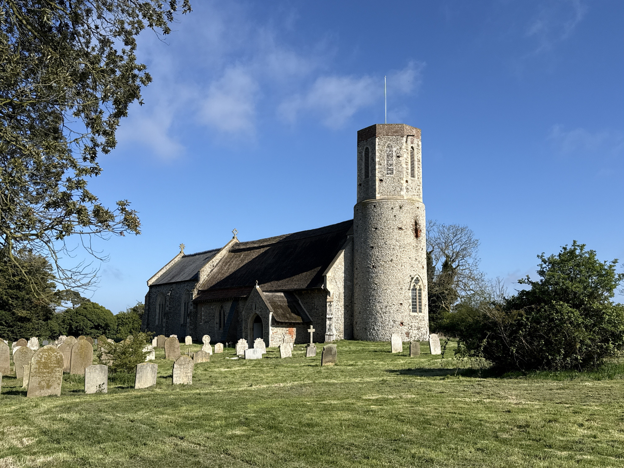 This media file captures a sunlit view of a historic English church and its surrounding graveyard, likely located near Great Yarmouth, United Kingdom. The central subject is an ancient church built primarily of light-colored stone, possibly flint, accented with reddish brickwork, particularly on its distinctive tower. The church features a unique round tower that transitions into an octagonal or hexagonal belfry at its apex, topped with a flagpole. Its roof is steeply pitched and appears to be covered with a dark, traditional material such as thatch or slate. Arched windows with simple tracery are visible on the church walls and tower.
The church is set within a verdant green churchyard, which is filled with numerous weathered gravestones of various shapes and sizes. These headstones, ranging from upright slabs to smaller markers, show signs of age with some visible moss or lichen, suggesting a long history of burials.
A large tree with branches and some foliage partially frames the upper left corner of the image. Additional trees and dense green bushes border the church grounds, adding to the tranquil, natural setting. The time of day appears to be morning or early afternoon, with bright, clear sunlight illuminating the scene and casting moderate shadows. The sky is a vibrant blue with a few wispy white clouds, contributing to the peaceful atmosphere. No specific activity or event is taking place, and no legible text is visible on the gravestones or the church. The overall scene evokes a sense of history and serenity.