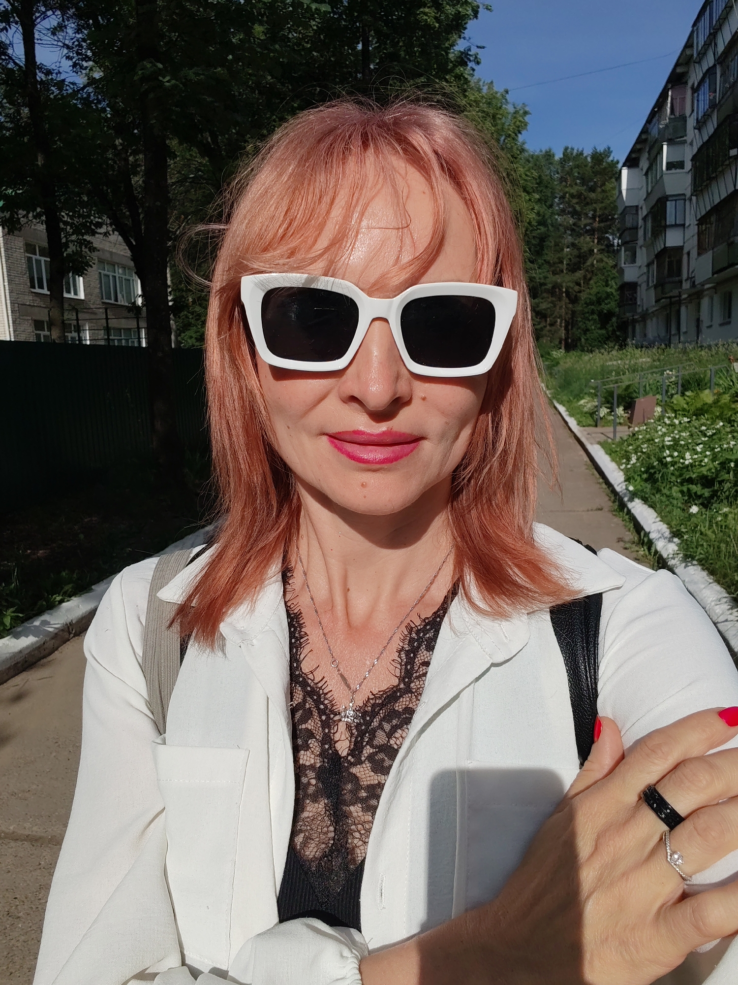 In Chaikovsky, Russia, a woman with pink hair and white sunglasses stands outdoors, posing for a selfie. She wears a white collared shirt over a black lace top, a silver necklace, and rings on her left hand. Her right hand is adorned with red nail polish. The background shows a paved walkway, green trees, and residential buildings.