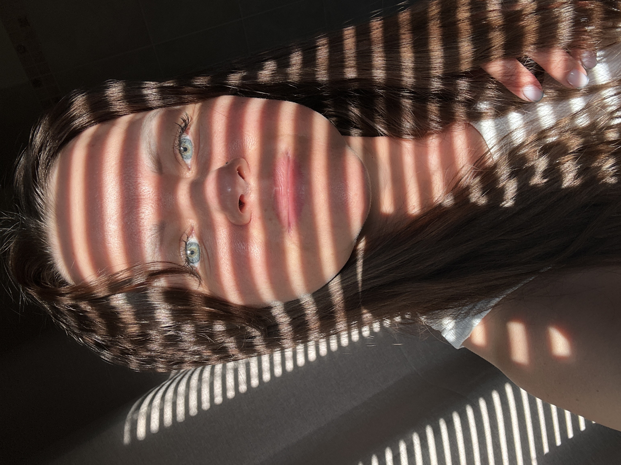 A close-up, indoor portrait depicts a young woman with brown hair and light blue eyes, likely in Chaikovsky, Russia. Her face is partially covered by strong, linear shadows, suggesting light filtering through window blinds. The woman's skin appears fair, and her lips are slightly parted. Her hair frames her face and is partially illuminated. Her hand rests near her head, showcasing pale fingernails. The background appears to be a solid-colored surface, likely a wall or bedding, also affected by the shadows. The overall orientation of the image suggests the subject is lying down.