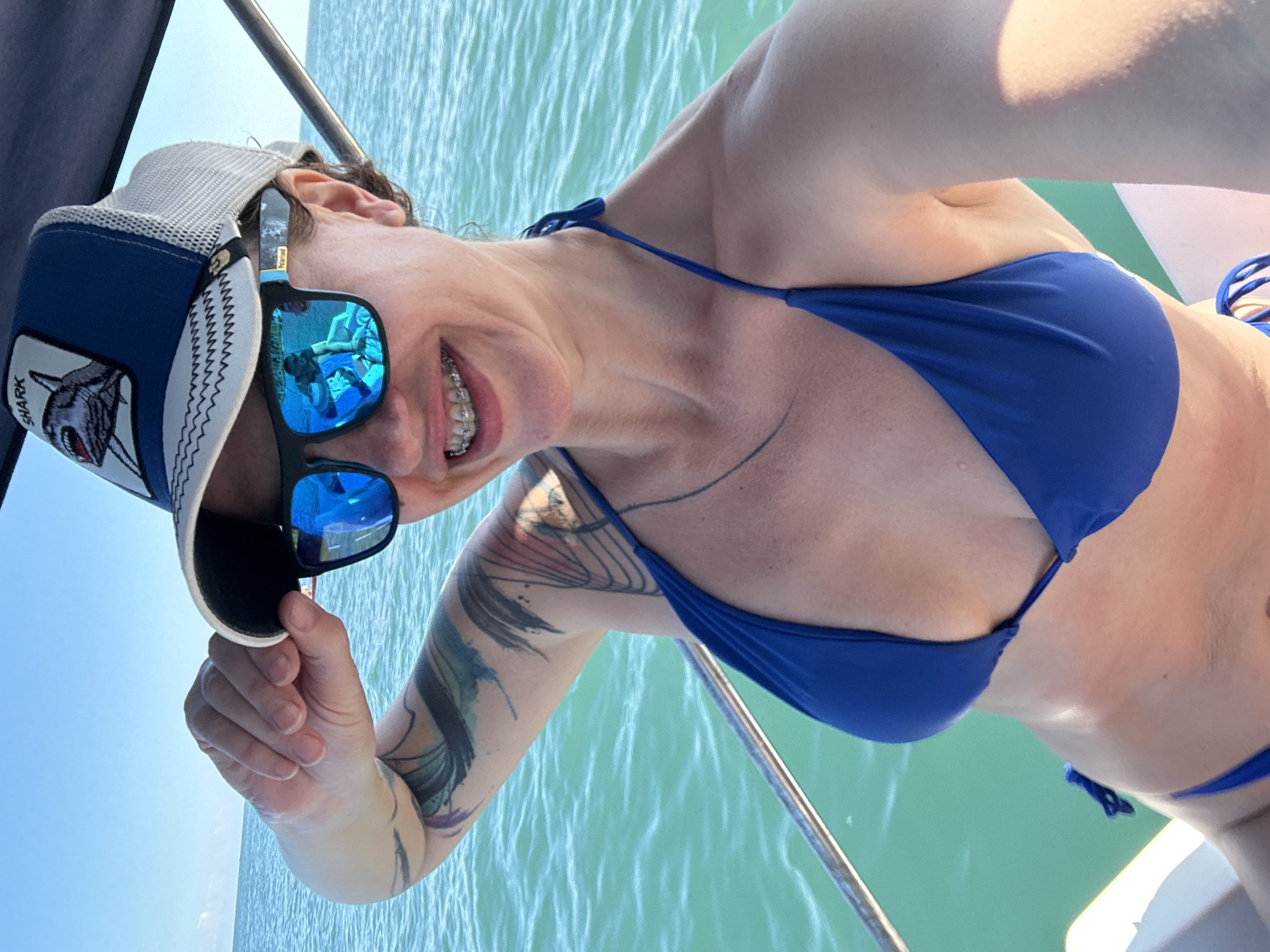 A woman is on a boat in the ocean, wearing a blue bikini and a blue trucker hat with a shark design. She is smiling and holding the brim of her hat. Her sunglasses reflect the sky and another person. The water is turquoise, and the sky is clear and blue. The image appears to be taken in a sunny, tropical or subtropical location.
