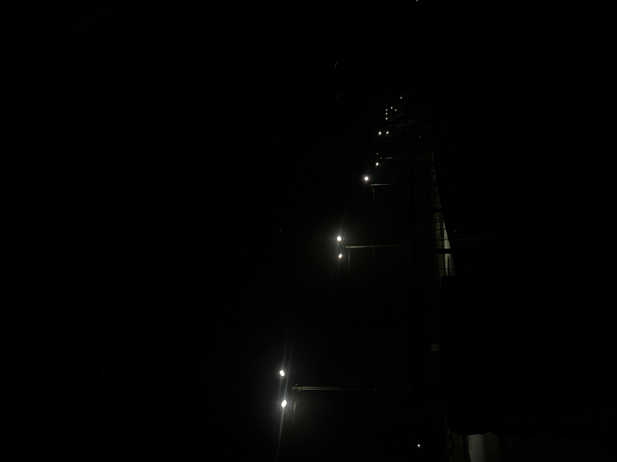 The image shows a dark night scene in what appears to be an industrial area of Galabovo, Bulgaria. A tower-like structure dominates the right side of the frame, with several lights illuminating its height. The lights are spaced at regular intervals, suggesting they might be mounted on supports or platforms attached to the tower. The surrounding environment is obscured by darkness, but some details of the structure's silhouette are visible. There is no clear indication of people or specific actions in the scene.