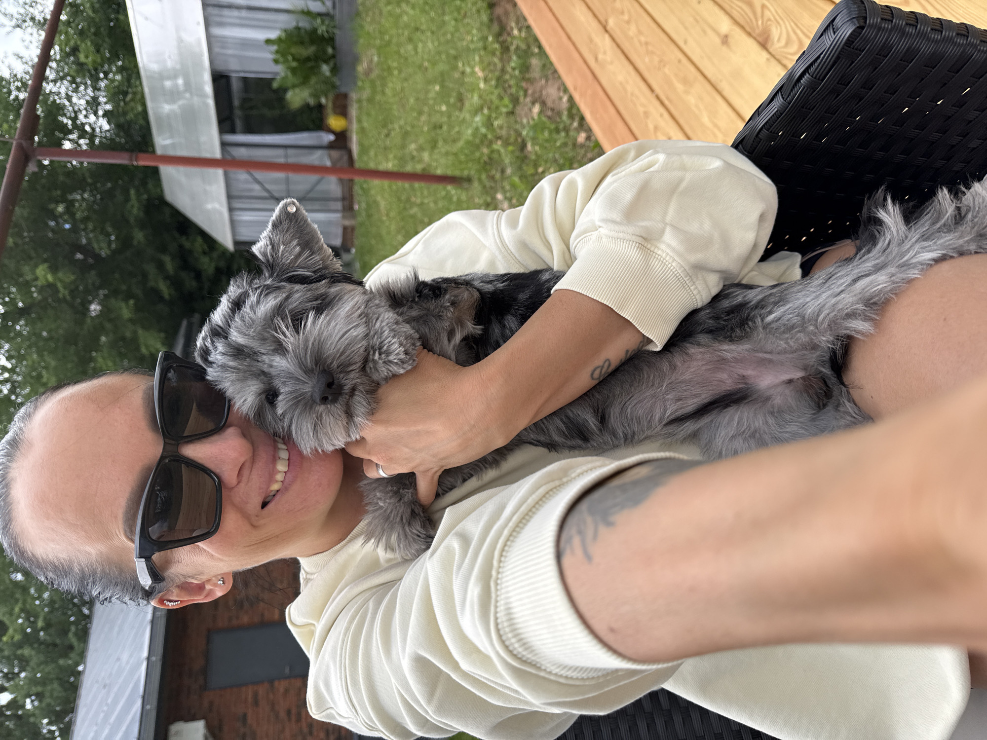 A woman wearing sunglasses and a light-colored sweatshirt is holding a small gray and black dog. The woman is smiling and looking at the camera, holding the dog close to her chest. The dog is looking towards the camera with its mouth slightly open. They are outdoors, with a wooden deck and a grassy area in the background. There are also some buildings and trees visible. The scene appears to be a casual, outdoor setting. The presence of a greenhouse structure suggests it might be a backyard or a garden area. The woman has tattoos on her arm and is wearing earrings. The dog appears to be a puppy with fluffy fur.