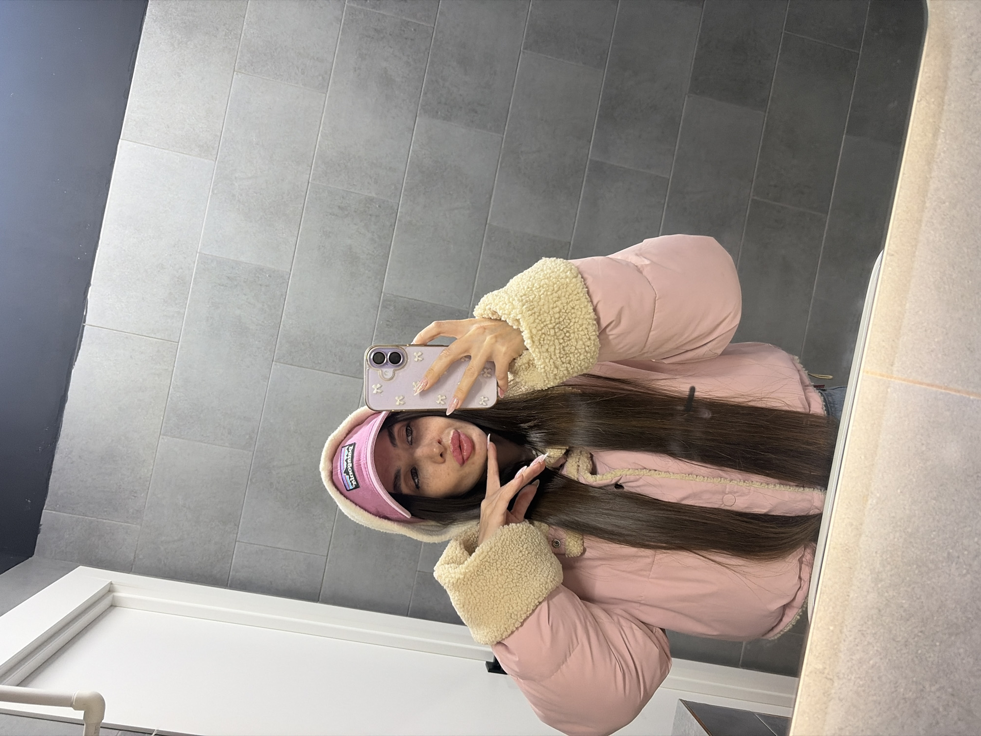 A woman is taking a selfie in what appears to be a bathroom. She is wearing a pink puffer jacket with a beige fur-lined collar and cuffs, and a pink baseball cap with a logo. She is holding a lavender-colored cell phone with a floral design. Her hair is long and dark brown. She is making a peace sign with her left hand. The background consists of gray tiled walls and a white door frame. The location is likely in Saint Petersburg, Russia, due to the gray color of the tiled walls.