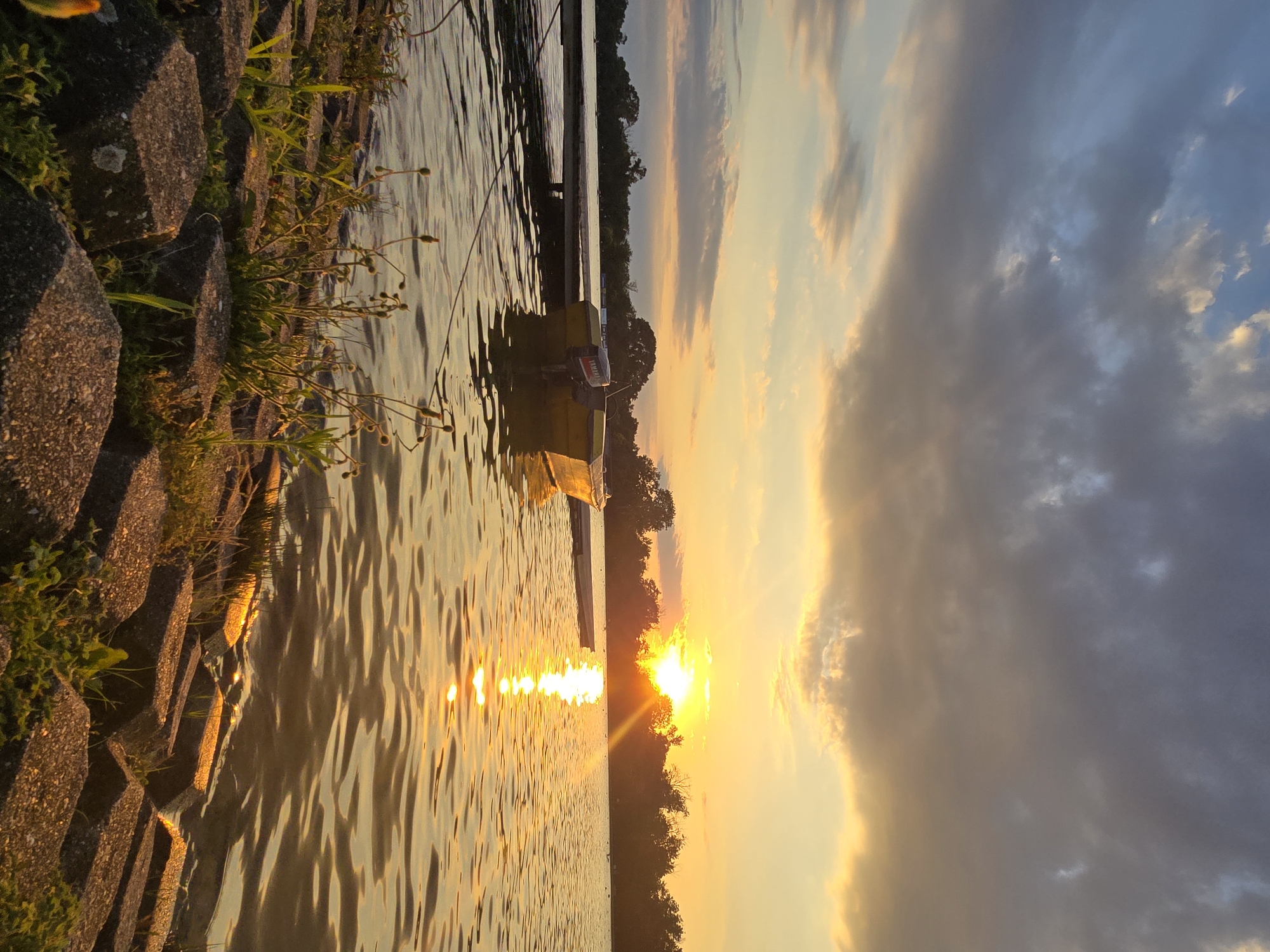 The image captures a serene sunset over a body of water, possibly a lake or a wide river. The sun, a bright orb, is setting on the horizon, casting a warm, golden glow across the scene. The sky is a canvas of colors, with dramatic clouds tinged with orange, yellow, and grey, suggesting a dynamic weather pattern. In the foreground, the water is rippled, reflecting the vibrant colors of the sky and the sun's descent.
Along the shoreline, a rough embankment made of dark, angular stones is visible, interspersed with patches of green grass and some small, dried plants with buds. A narrow wooden pier extends into the water, and a small, yellow boat with a Yamaha outboard motor is moored to it. The boat appears to be empty. In the background, a line of trees forms the distant shore, silhouetted against the bright sky.
The overall atmosphere is peaceful and tranquil, evoking a sense of calm as day transitions into evening. There are no people visible in the frame, and the scene is devoid of any obvious activity, focusing solely on the natural beauty of the sunset and its reflections. The image appears to be a landscape photograph, likely taken from a waterfront location.
Without specific visual cues linking it to Plovdiv, Bulgaria, it is impossible to confirm the location. However, if this scene were to be in Plovdiv, it could potentially be along the Maritsa River, which flows through the city, or perhaps a smaller lake or reservoir within or on the outskirts of the urban area. Cities often have waterfront parks or recreational areas that could resemble this setting.