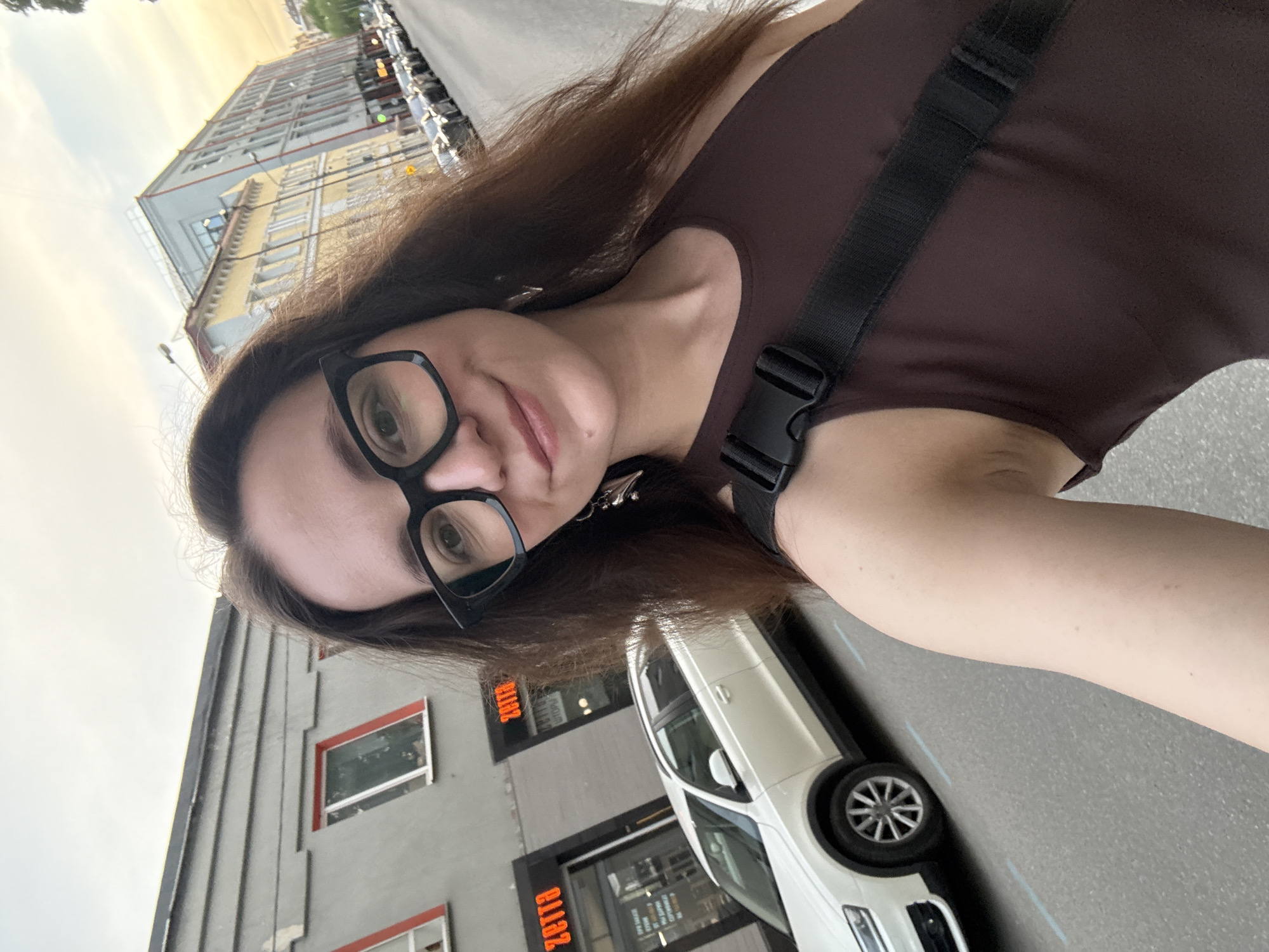 A woman with dark hair and glasses is taking a selfie outside in Moscow, Russia. She is wearing a brown top with a black strap across her chest. Behind her is a white car parked on the street, and a building with "Zetta" written in reverse order. In the background, other buildings are visible. The sky appears to be overcast.