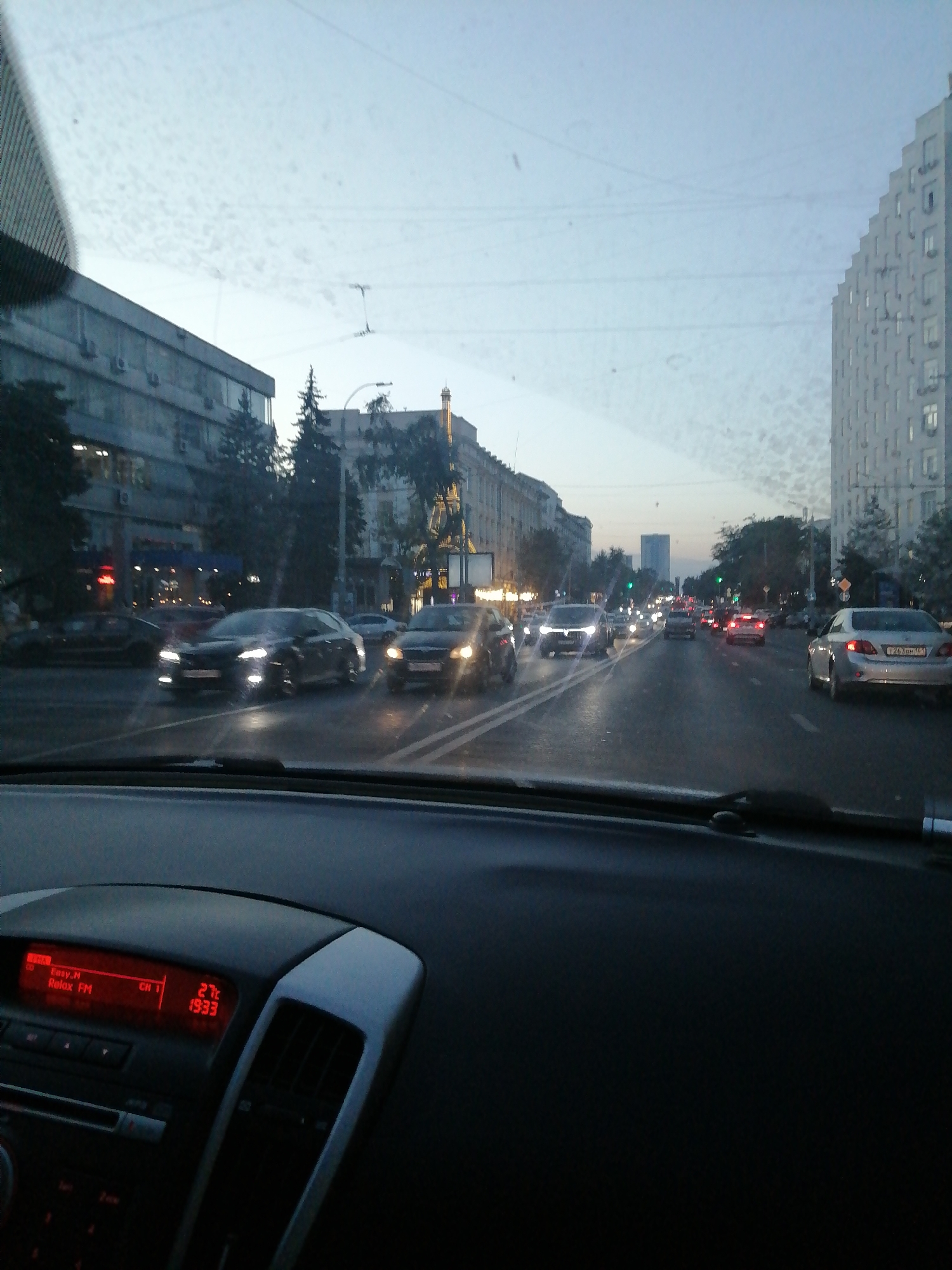 The image is a point-of-view shot from inside a car, looking through the windshield. The time displayed on the car's dashboard is 19:33, and the temperature is 27 degrees Celsius. The car is in traffic on a multi-lane street in Rostov-on-Don, Russia. The street is lined with buildings on both sides, including a tall, modern-looking building on the right. The sky is overcast and it appears to be dusk. There are numerous cars on the road, some with their headlights on. The traffic appears to be moderate to heavy. The scene depicts urban street life during the evening commute.