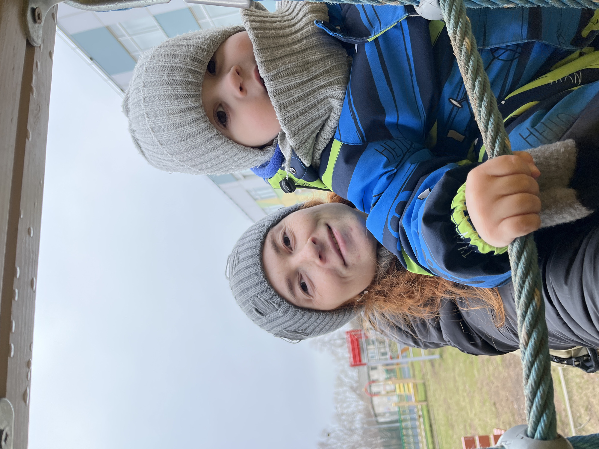 An adult and a child are captured from a low-angle perspective, looking downwards at the camera. Both are dressed in warm winter attire, suggesting cool or cold weather conditions. The adult, with auburn hair, is positioned in the foreground, wearing a grey knitted beanie and a black jacket, and has a gentle smile. Behind and slightly above the adult is the child, also sporting a grey knitted beanie that covers their ears and neck, and a blue and green striped winter jacket with faint "HELLO" text visible on it. The child looks into the camera with wide, curious eyes.
Both individuals are holding onto a thick, blue-green braided rope, which appears to be part of a playground climbing structure. A blurred wooden beam is partially visible on the left side of the frame. The background consists of a bright, overcast sky, indicative of a cloudy day with diffused light. Below the subjects, the out-of-focus background reveals elements of a colorful outdoor playground, including red, yellow, and blue structures and patches of grass. The scene depicts a moment of play or interaction at a playground in Chaikovsky, Russia.