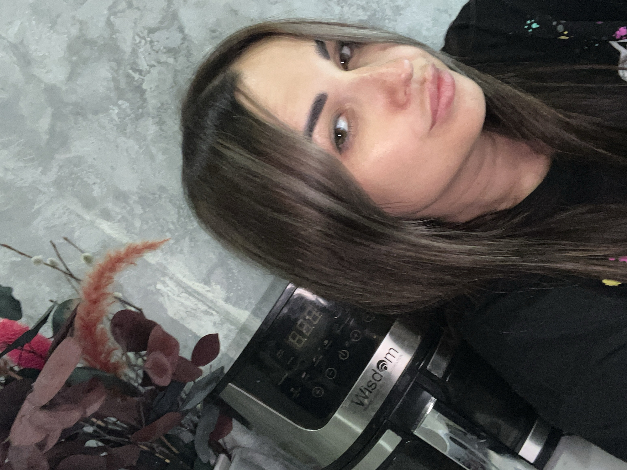 A woman is taking a selfie in an indoor setting. She has long, dark brown hair with highlights, and is wearing a black t-shirt. Her face is partially visible, and she is looking at the camera with a slight smile. To her left, partially obscured by her hair, is a kitchen appliance, possibly an air fryer, with a digital display showing "888". Behind her, there is a bouquet of dried flowers and plants. The wall behind her has a textured, grey concrete finish. The location is identified as Unknown, Unknown.
