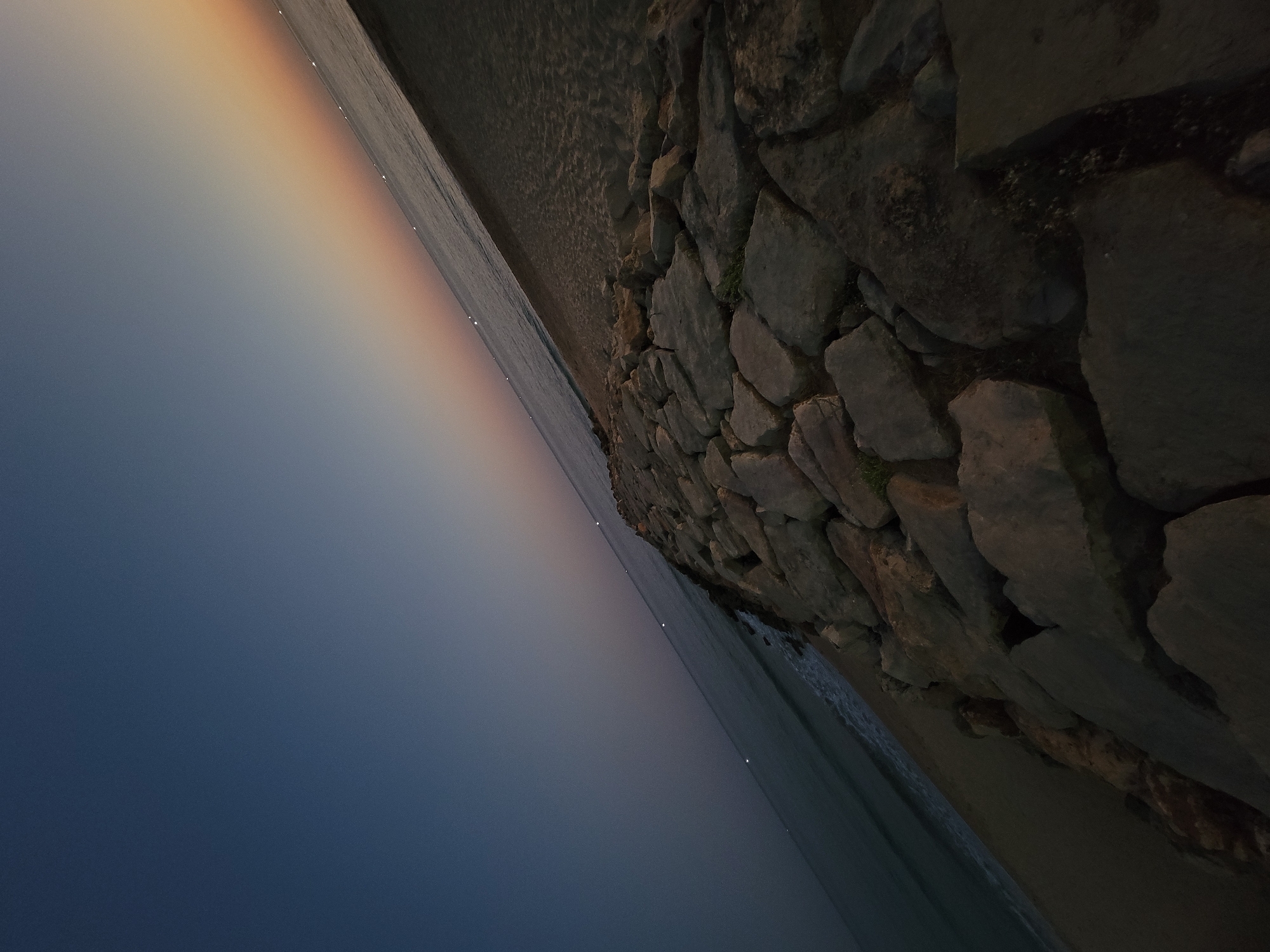 The image depicts a serene coastal scene at Quarteira, Portugal, likely during twilight or dawn. The foreground is dominated by a man-made stone wall, constructed from large, irregularly shaped rocks, creating a rugged texture and a sense of permanence against the natural elements. Small patches of green vegetation are visible growing between some of the stones, suggesting a slightly damp or fertile environment.
Beyond the wall, a sandy beach stretches out, meeting the calm sea. The water is a deep, muted blue, reflecting the dusky sky. A subtle, diffused band of light, exhibiting colors ranging from soft orange and yellow to a pale pink and lavender, stretches across the horizon where the sky meets the sea. This phenomenon could be a remnant of a sunset or the anticipation of a sunrise, casting a gentle glow over the water.
Dotted along the distant waterline, a series of small, bright white lights are visible. These lights could indicate buoys, distant boats, or even lights from structures further along the coast, adding a touch of human presence to the otherwise natural and tranquil landscape.
The overall mood of the image is peaceful and contemplative. The low light conditions and the gentle lapping of the waves contribute to a sense of calm. The perspective, angled slightly upwards from the base of the stone wall, emphasizes the vastness of the sky and sea. No people or specific text are visible in the image. The location is identified as Quarteira, Portugal.