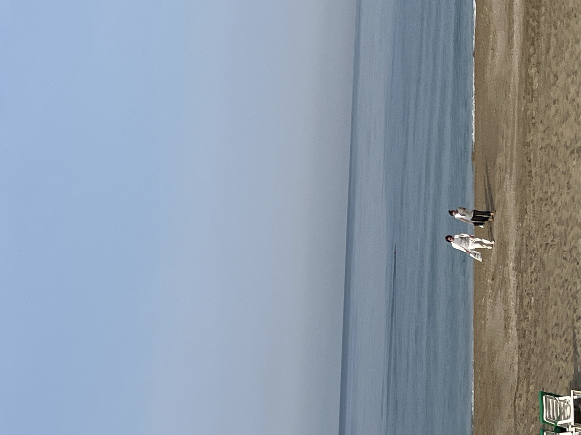 The image depicts a beach scene in Alanya, Turkey. Two people stand on the sand near the water's edge. The sky is clear and blue, and the sea is a gradient of blue hues. A few beach chairs are visible in the lower right corner.