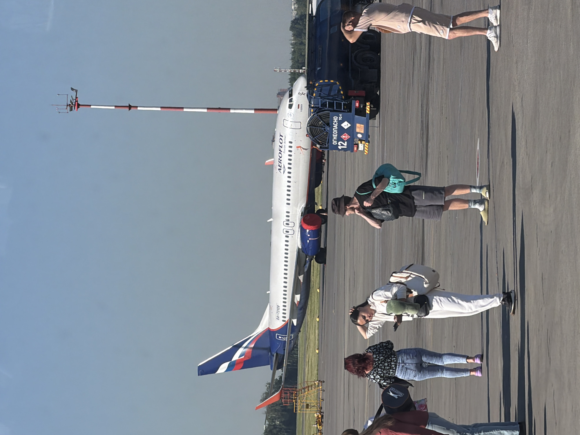 This image depicts a scene at an airport, likely Sheremetyevo International Airport in Khimki, Russia, given the Aeroflot aircraft. In the background, a white Aeroflot airplane with the registration RA-73109 is parked on the tarmac. To the right of the aircraft, a blue fuel truck with "ОГНЕОПАCНО" (Flammable) signage is visible. In the foreground and midground, several individuals are standing on the tarmac, some carrying bags and appearing to be waiting. One person in a white outfit is holding a phone and a green object. Another person in a black t-shirt and shorts is holding a black bag and a teal bag. The sky is clear and blue, suggesting a sunny day. The ground is asphalt, typical of an airport apron. A red and white striped weather tower is situated in the background.