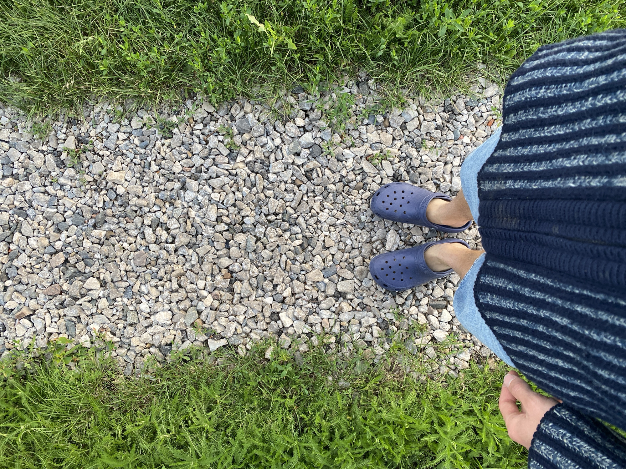 A top-down view shows a person standing on a gravel path bordered by green grass. The individual is wearing blue Crocs and jeans, with a dark blue striped sweater draped over their legs. Their left hand is visible near the bottom right corner of the frame, resting on their lap. The scene appears to be outdoors, with natural lighting suggesting daytime. The environment suggests a casual outdoor setting, possibly a park or a backyard. No text is visible in the image.