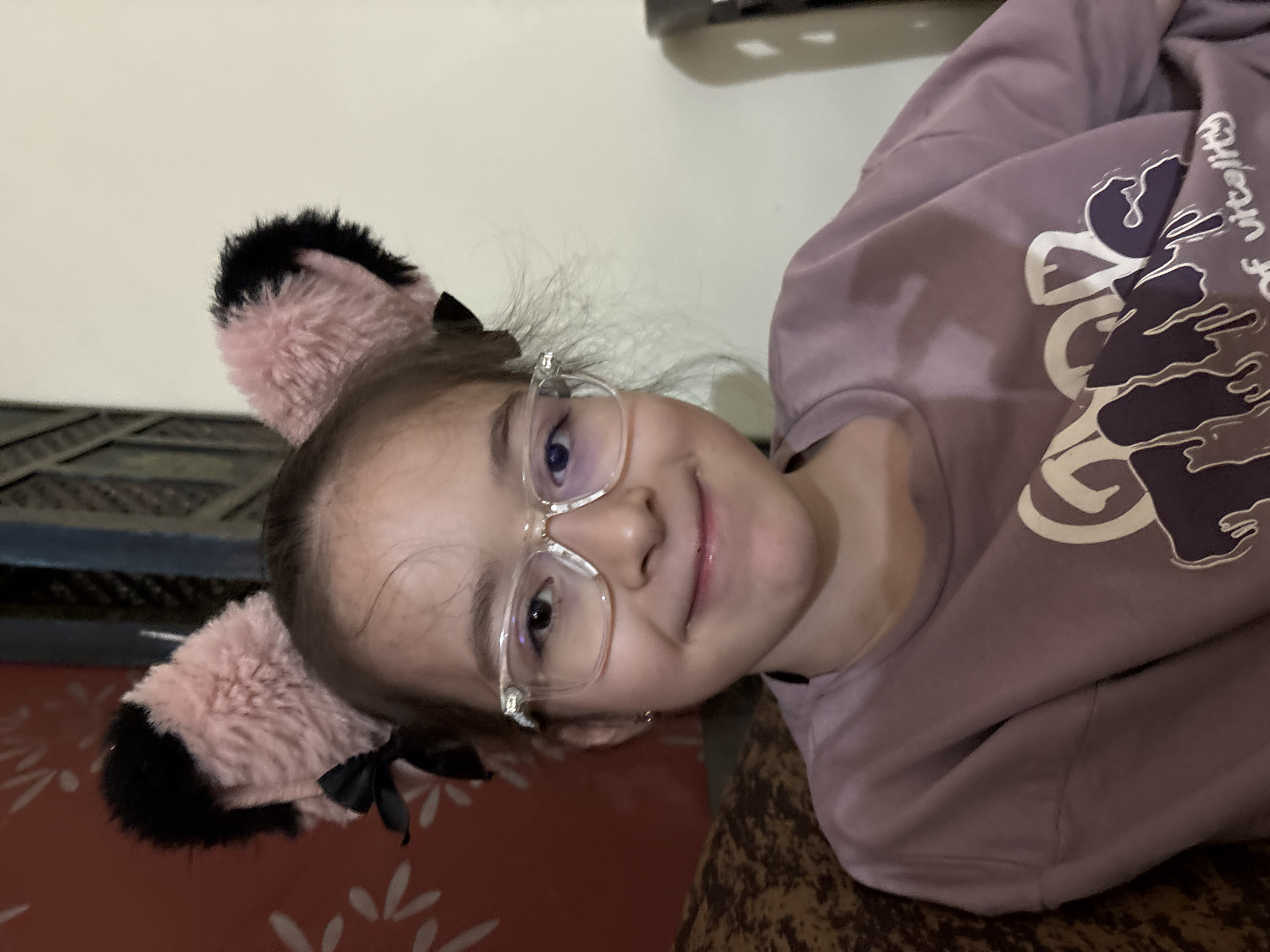 A young girl with brown hair, wearing clear-rimmed glasses and a mauve sweatshirt, smiles directly at the camera. She has pink and black cat ears with small black bows attached to them, perched on her head. The background shows a red, patterned wallpaper on the left and a plain, light-colored wall on the right. The girl is seated on what appears to be a couch. The context of Ashvem, India, does not appear to be depicted in the visual elements of the image.