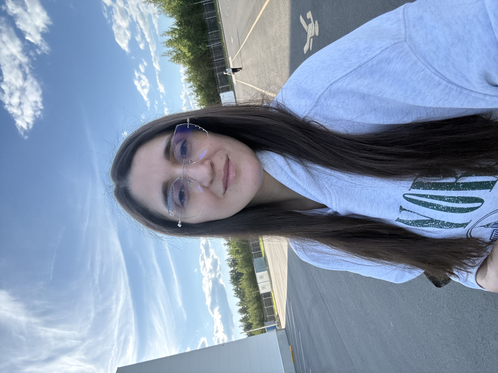 A woman with long, dark brown hair, wearing rimless eyeglasses and a light grey hooded sweatshirt with a partial green graphic print, is depicted in a self-portrait, smiling faintly. The scene is outdoors on a grey asphalt surface, marked with yellow lines and a white stylized airplane symbol in the lower right foreground, indicative of an airfield or paved aviation area. In the far distance on the asphalt, a lone person walks away from the camera. To the left, a tall, white, conical structure stands alongside a white building or wall. A dense line of evergreen trees forms a backdrop across the mid-ground, located behind fences and several small, rectangular utility buildings. The upper portion of the image shows a blue sky with scattered white cirrus and cumulus clouds. The visible elements, particularly the airfield markings, align with an outdoor location in Sarapul, Russia, as specified.