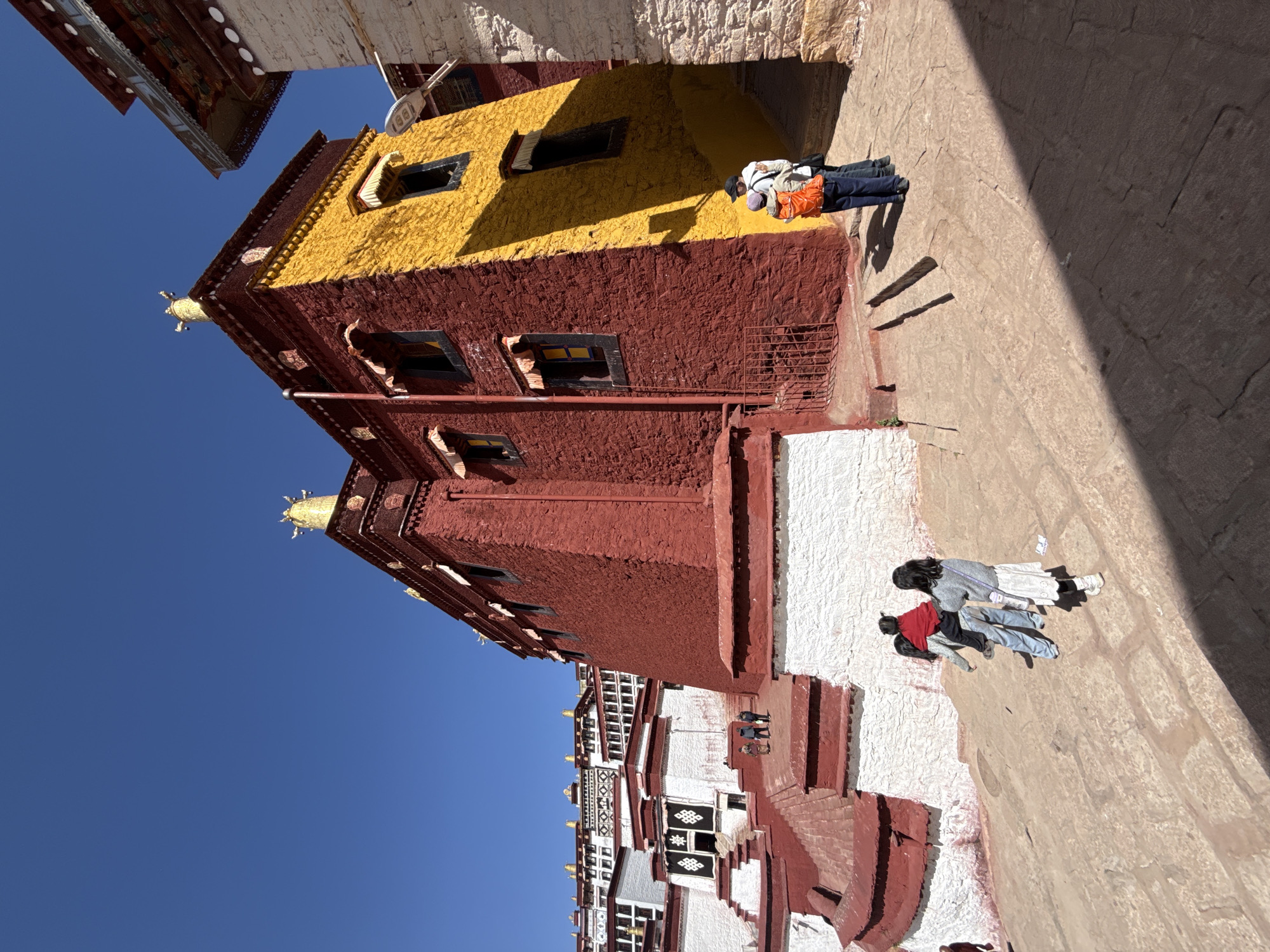 The image shows a building in Lhasa, China, likely part of the Potala Palace complex, a prominent Tibetan Buddhist monastery and former residence of the Dalai Lama.
The architecture features a multi-tiered structure, characterized by the distinctive red and yellow facades, and white-washed walls. People are shown walking outside of the building. The sky is visible as a clear blue.