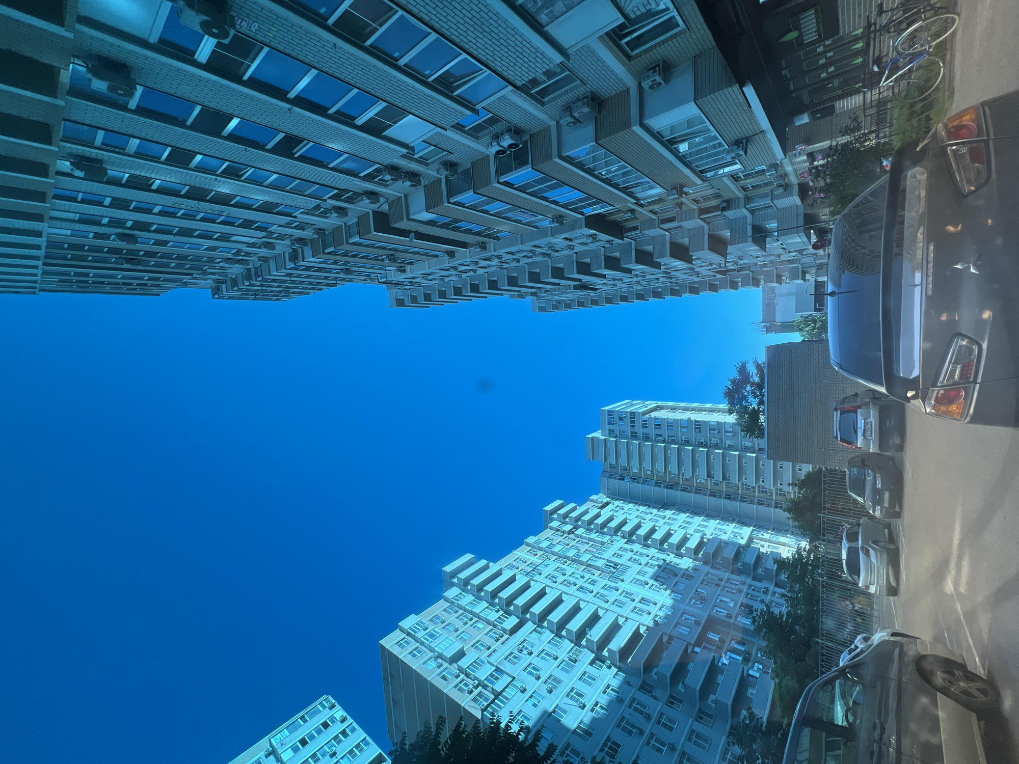 The image depicts a low-angle view of a modern urban residential environment in Krasnodar, Russia, specifically within the "Novy Gorod" residential complex, likely near 148/2 Ulitsa Turgeneva. The scene is dominated by a clear, vibrant blue sky filling the upper half of the frame.
Multiple high-rise residential buildings, predominantly light-colored with brick and stucco facades, frame the sky. The building on the right is tall and close, extending beyond the top of the frame, featuring numerous recessed balconies and a dense arrangement of external air conditioning units. Its facade shows alternating light and darker brown brick sections. On the left, another large residential building stands further back, also with many balconies and AC units, partially obscured by green trees. A third, partially visible building is present on the far left.
The photograph appears to be taken from inside a vehicle, evidenced by reflections and a slight blue tint across the buildings, likely from a tinted windshield. In the foreground, a paved parking lot is visible with white demarcation lines. Several cars are present:
* Partially visible on the far left, a dark-colored minivan or SUV.
* A silver Opel Astra H hatchback (Russian license plate `O588HP 196`) parked in the middle-left.
* A dark-colored sedan, possibly a Lada Priora, parked behind the hatchback.
* A silver Nissan X-Trail T30 SUV (Russian license plate `T317TC 93`) parked in the middle-right.
* The rear of a dark-colored Mitsubishi Lancer X sedan occupies the immediate foreground on the right.
A metal fence separates the parking area from a small patch of trees and a low-rise structure in the mid-distance. On the far right, partially visible behind the Mitsubishi Lancer, is the dark facade of a ground-floor commercial establishment or shop, with several bicycles parked against its wall. No people are discernible in the scene. The overall impression is a sunny day in a densely populated, modern residential district.