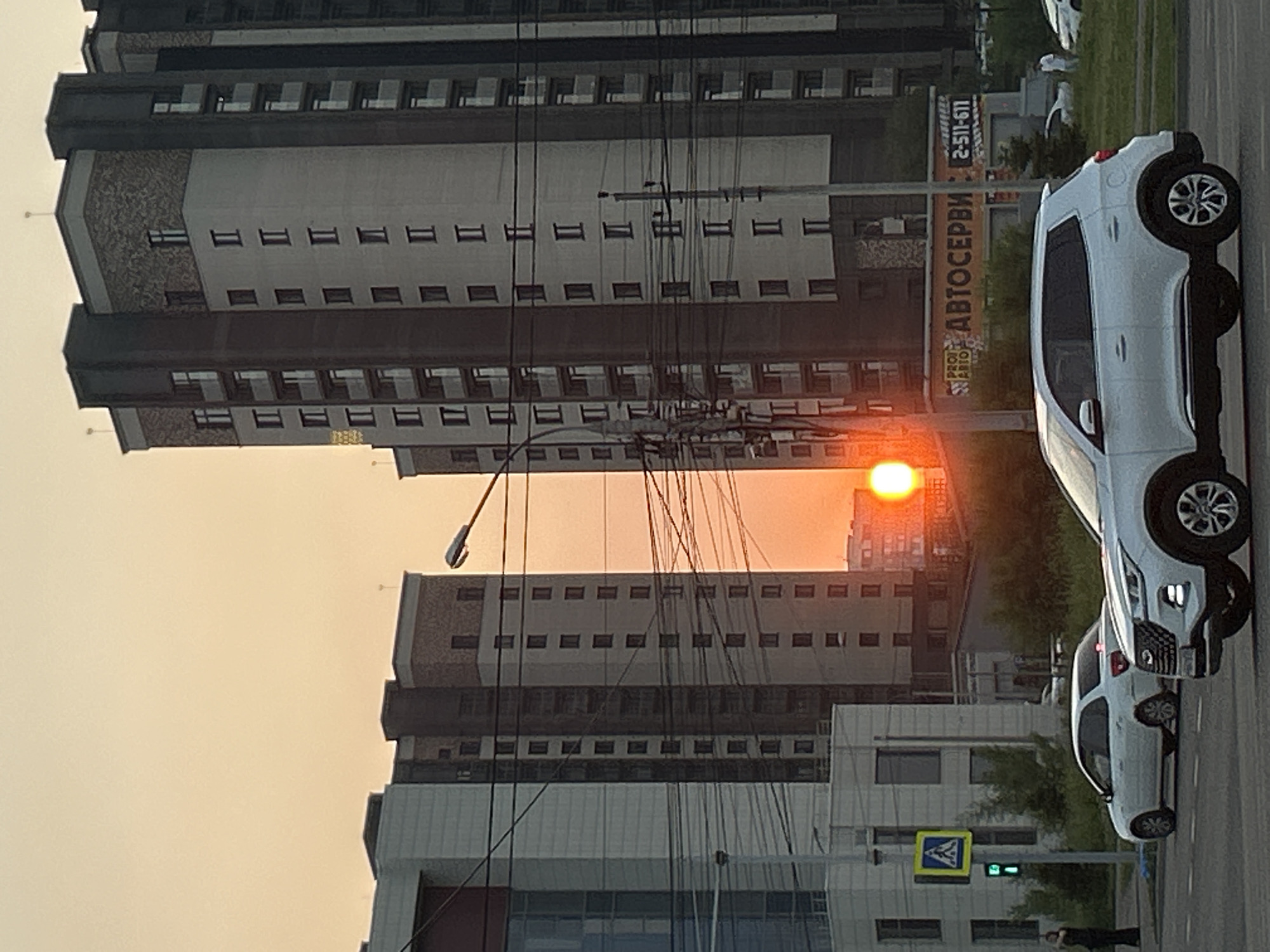 This media file shows a street scene at sunset in Krasnoyarsk, Russia. The foreground features two white SUVs parked on the right side of the road. The first SUV, a larger model, is parked further down the street, while a second, smaller SUV is closer to the viewer. A pedestrian is visible standing near the second SUV.
In the background, several multi-story buildings rise into the sky. These buildings are a mix of modern and older architectural styles, with visible windows and balconies. A prominent yellow billboard with Russian text is attached to one of the buildings. The text on the billboard is partially obscured but includes Cyrillic letters and what appears to be a phone number: "АВТОСЕРВИС 2-511-671".
The sun is setting, casting a warm, orange glow across the sky and reflecting off the buildings. The light suggests it is late evening. The weather appears to be clear, with no visible precipitation. The scene is urban, characterized by the presence of buildings, vehicles, and a road.