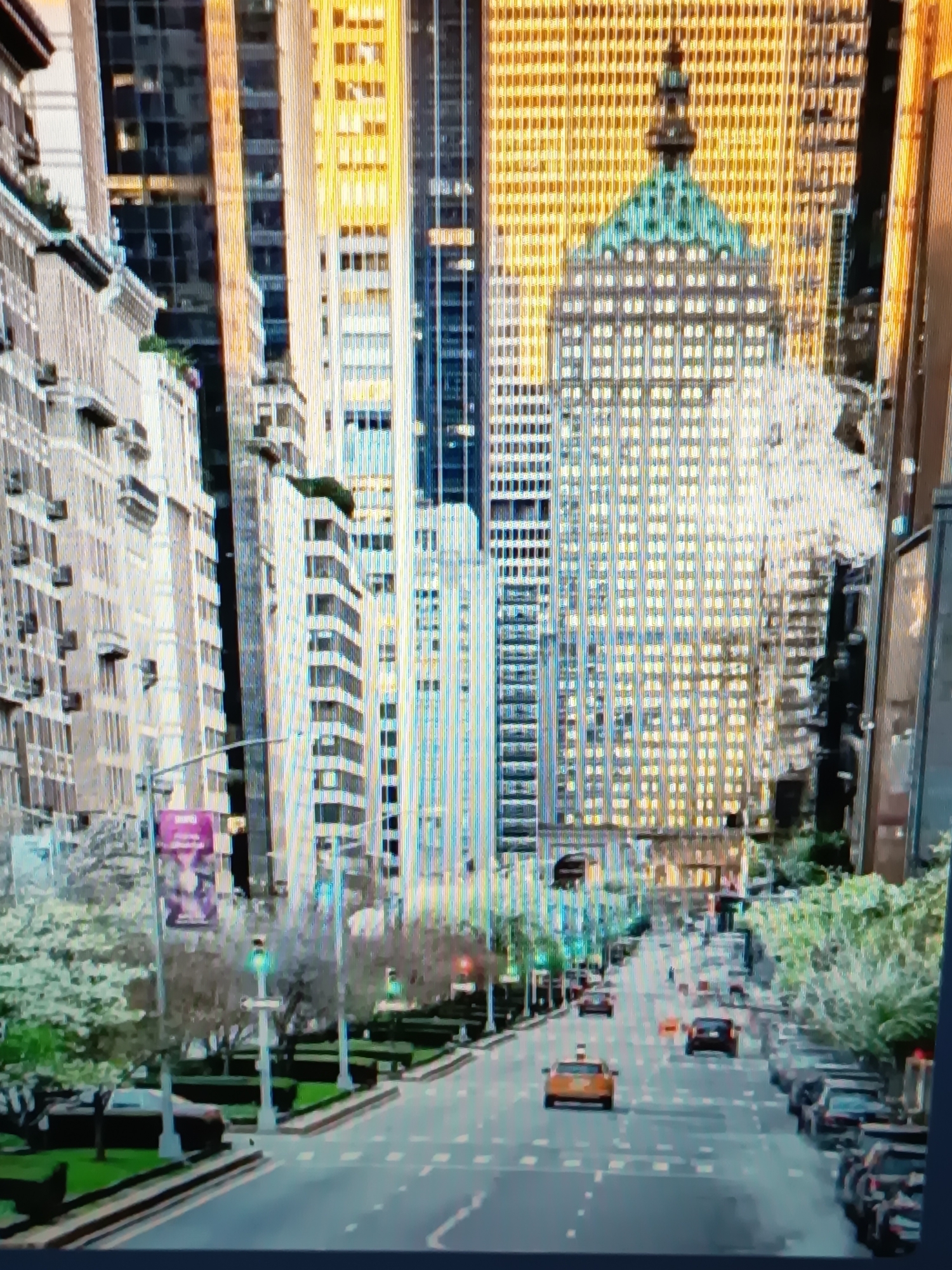 The image depicts a city street scene, likely in a major metropolitan area, given the presence of numerous tall buildings with a significant number of windows. The architecture suggests a commercial or residential district. The street itself is paved and has lane markings, indicating it is a public roadway. Several vehicles are visible on the street, including a yellow taxi, a dark colored SUV, and other cars parked along the side of the road. Trees and landscaping are present along the sidewalks, suggesting a well-maintained urban environment. Traffic lights are visible, indicating traffic control measures. A large billboard or advertisement is mounted on a pole on the left side of the street. The overall impression is a bustling city during daytime, with buildings reflecting sunlight and a clear sky. The location is not Serhiivka, Ukraine, as the architecture and urban planning are not consistent with that region.