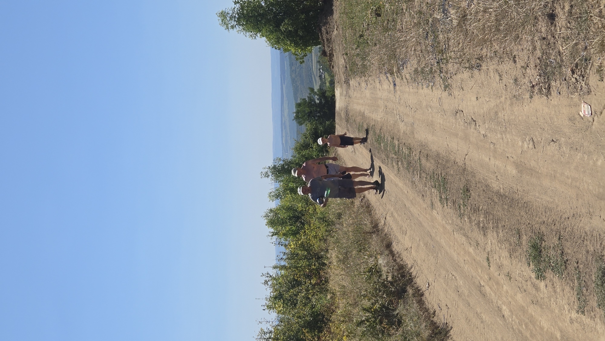Three individuals, two adult males and one young male, walk along a dirt road in a rural, hilly landscape under a clear blue sky. The two adult males are in the foreground, with the taller, shirtless man on the right and a man wearing a t-shirt and hat to his left. The younger male is walking behind them to the right. The road is dry and dusty, with sparse vegetation on either side, including dry grasses and scattered bushes and trees. In the background, a hazy, expansive view of rolling hills and distant greenery suggests a rural or semi-rural setting. The overall scene depicts a casual outdoor activity, possibly a walk or hike, in a natural environment. The location is identified as Bălănești, Moldova.