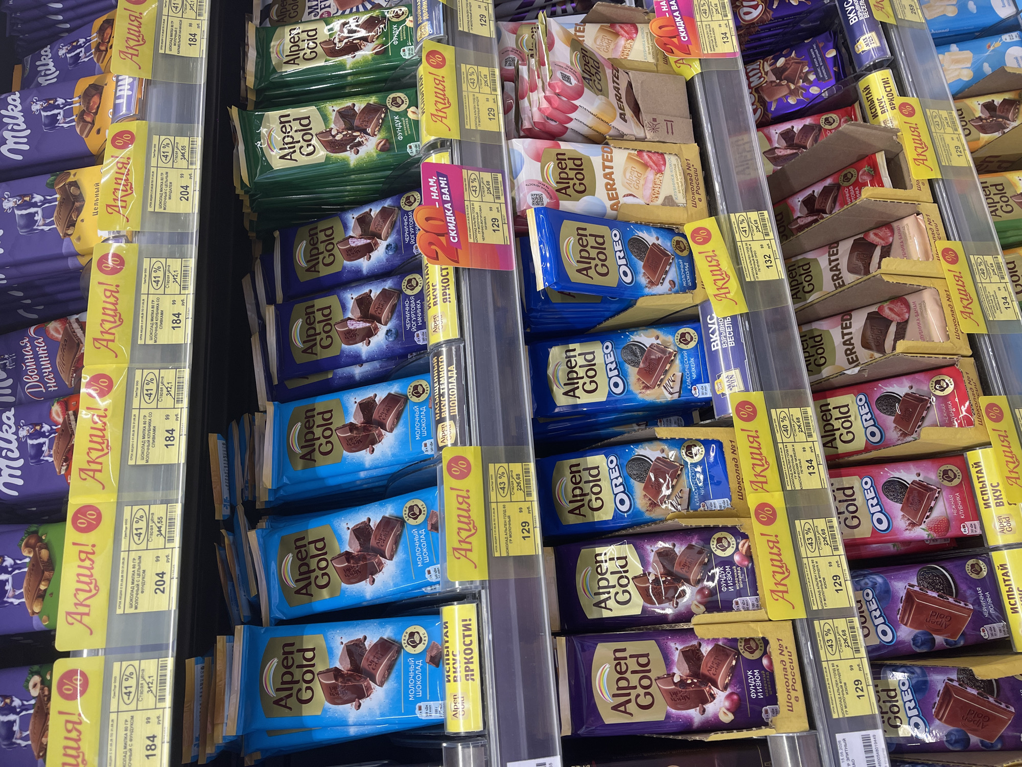 The image shows a supermarket shelf stocked with various Alpen Gold and Milka chocolate bars. The Alpen Gold bars come in different flavors, including milk chocolate, dark chocolate, Oreo, and fruit combinations. Several bars have yellow "Aktsiya!" (promotion) signs indicating discounts. Prices are listed in rubles, with both the discounted price and the original price displayed. The Milka bars also have similar "Aktsiya!" signs and discounted prices.