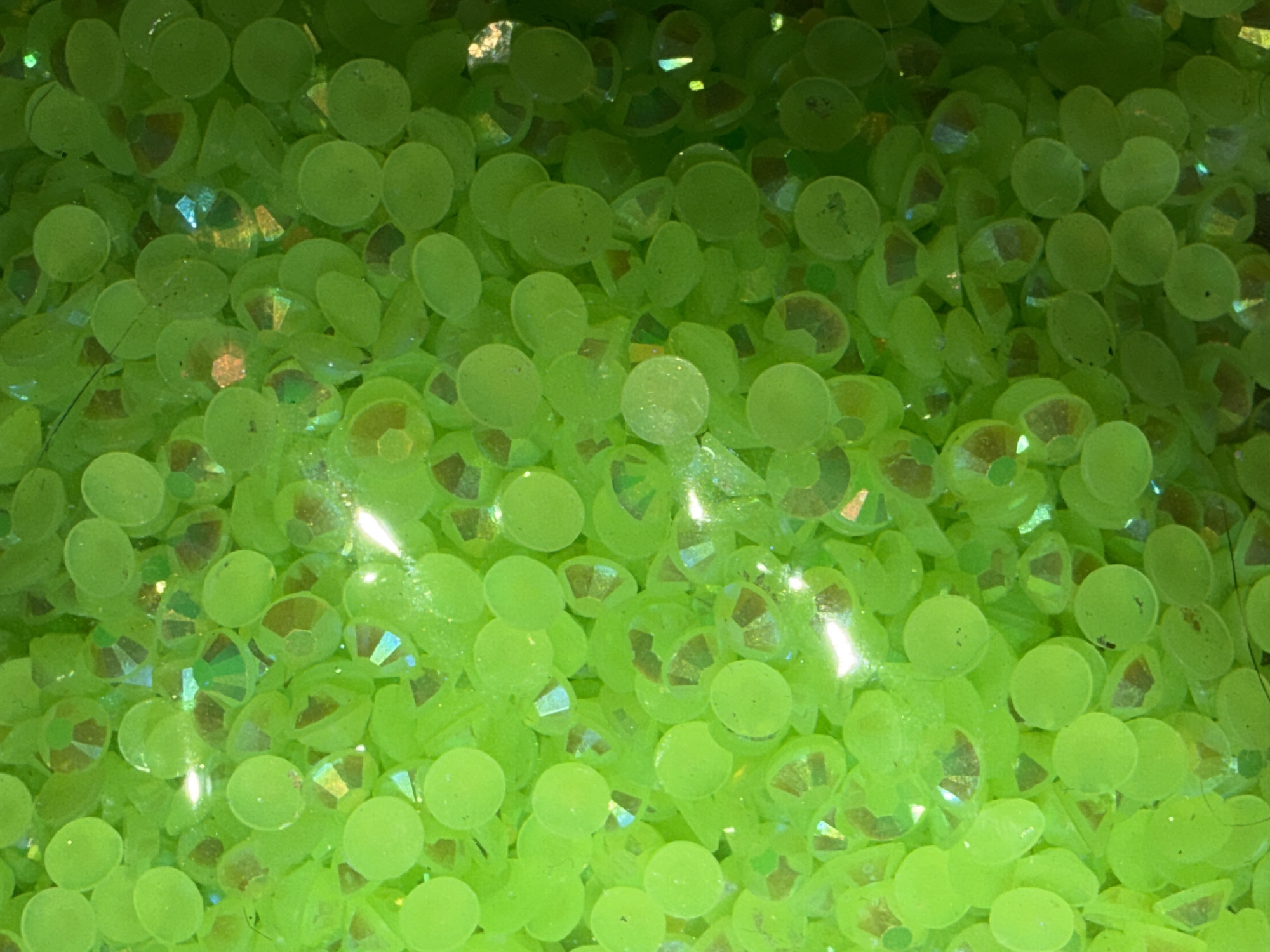The image is a close-up, macro shot of a pile of small, round, lime green rhinestones. Some of the rhinestones appear to be faceted, reflecting light with iridescent hues of yellow, orange, and blue. The overall impression is one of glitter and sparkle. The background is a gradient of dark green, suggesting a shadow or a deeper pile of rhinestones. The scene is not in a specific location in the city of Portland, United States of America, but rather appears to be a collection of craft supplies. There are no people, actions, or interactions depicted.