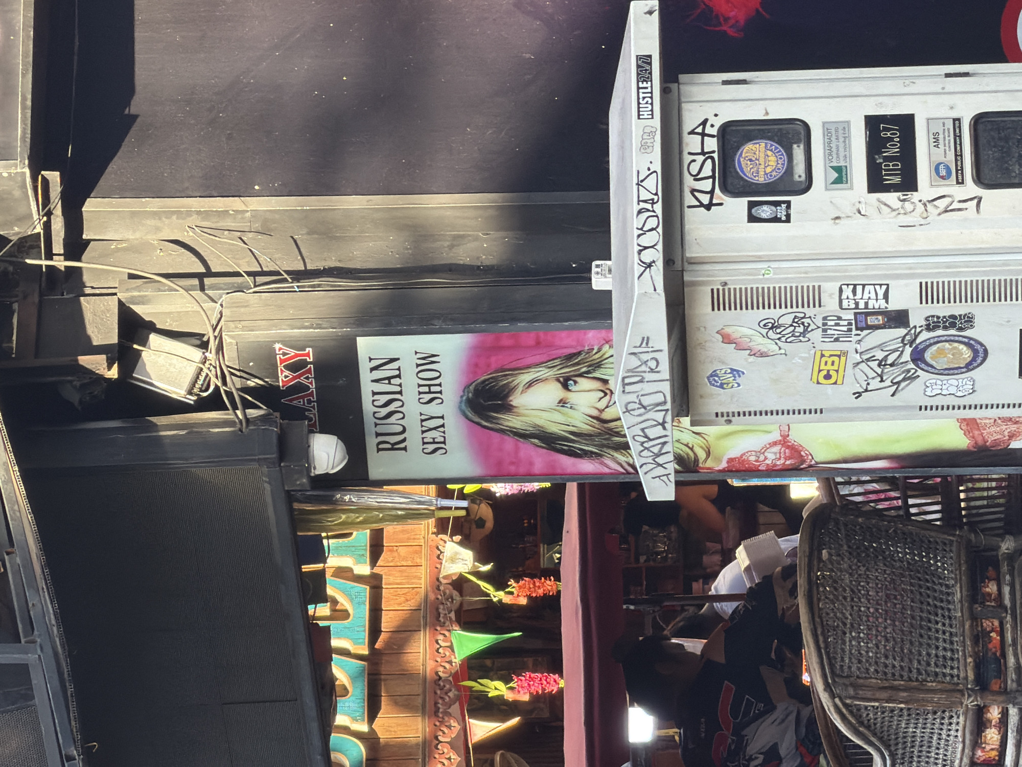 The image depicts a street scene in Pattaya City, Thailand, likely a commercial area with adult entertainment venues. In the foreground, a person is seated in a wicker chair. Behind them, a sign advertises "RUSSIAN SEXY SHOW" with a blonde woman's image. To the right of the sign is a white electrical box covered in stickers and graffiti. The background reveals more of the establishment's facade with wooden paneling and colorful decorations, suggesting a vibrant and possibly bustling atmosphere. The overall impression is one of a typical tourist-oriented entertainment district.