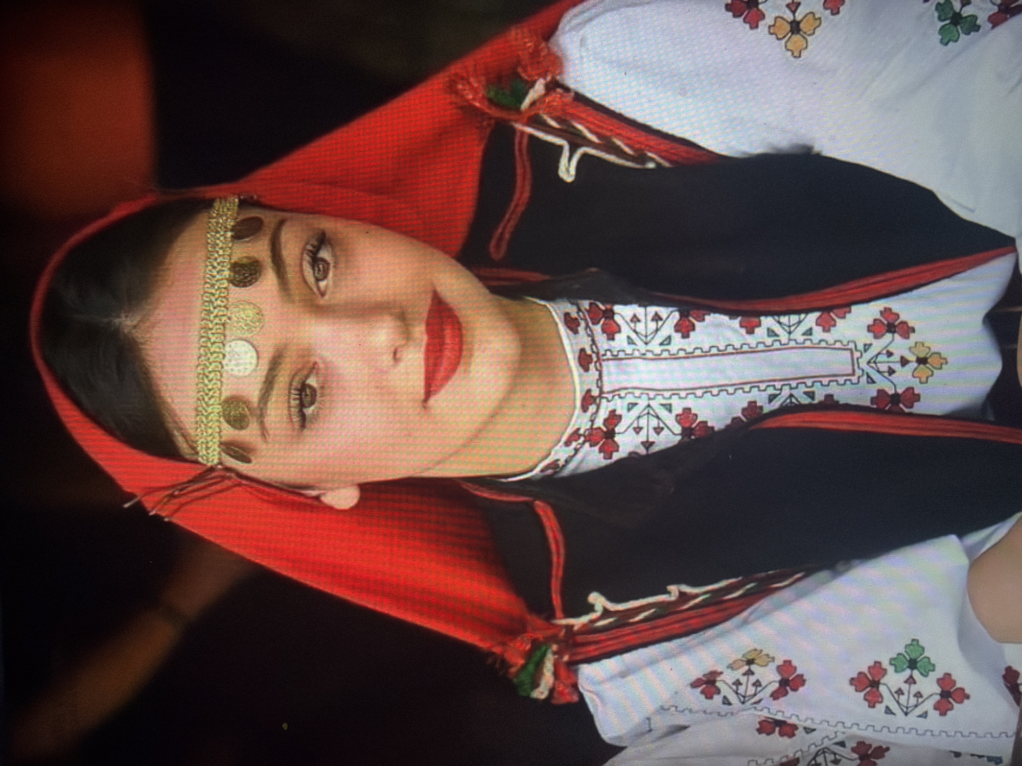A young female subject is depicted in a head-and-torso portrait, facing forward and looking directly at the viewer. She is dressed in traditional Bulgarian folk attire. Her head is covered by a vibrant red fabric that drapes over her shoulders and back. Across her forehead, she wears a gold-colored headband embellished with a series of approximately nine circular metallic discs, resembling coins. Her dark hair is visible beneath the head covering, parting in the center.
She has fair skin, dark brown eyes, and her lips are accentuated with bright red lipstick, forming a gentle smile. She wears a white long-sleeved garment, likely a blouse or shirt, which features intricate embroidery. The embroidery patterns include repeating red, green, and yellow floral motifs on the sleeves and scattered on the main body of the garment, as well as a more structured geometric and floral design around the neckline. Over this white garment, she wears a dark, likely black, vest or jacket that is trimmed with red piping along the edges and features decorative elements, including red, green, and white tassels or ribbons, on her left shoulder.
The background is dark and largely out of focus, with hints of warmer, reddish-brown tones in the upper left, suggesting an indoor or dimly lit environment. Portions of another person or object are partially visible on the far left of the frame. This depiction is associated with the city of Shumen, Bulgaria.