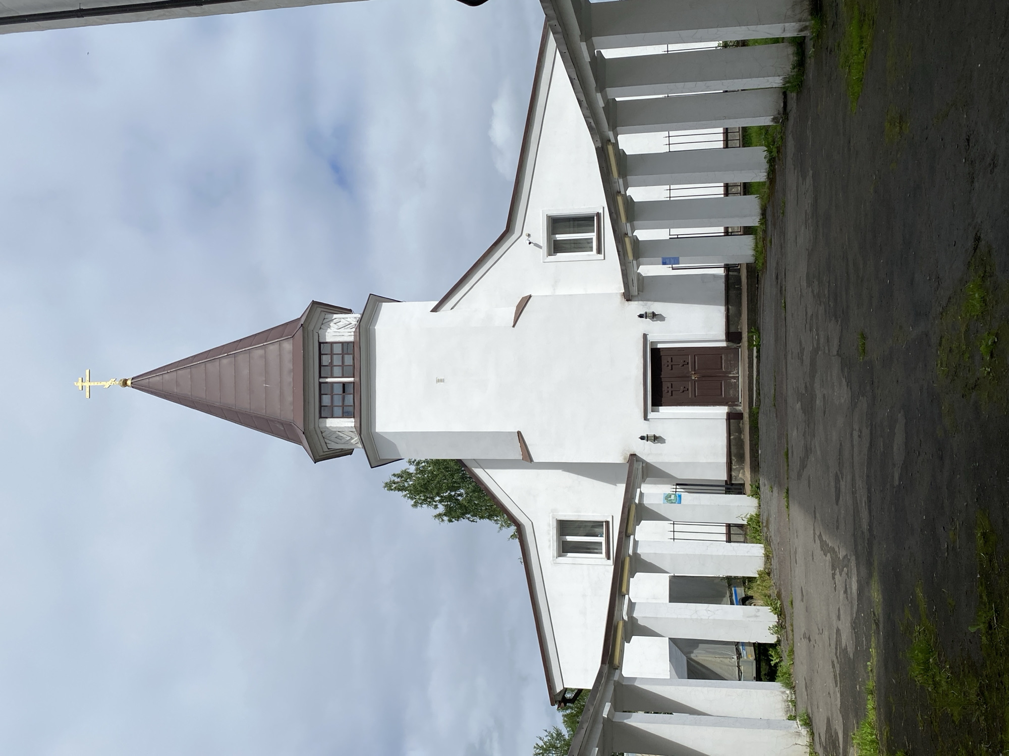 A frontal, slightly low-angle, eye-level shot captures the exterior of a white church in Sortavala, Russia. The church features a prominent, brown, spire-like roof topped with a golden cross. A rectangular section below the spire has a band of windows. The main body of the church is white with a brown-trimmed, steeply pitched roofline. There are two rectangular windows on the main facade, one on each side, and a set of double brown doors in the center. Flanking the entrance is a white colonnade with square pillars and a brown roof extending outwards. The ground in front of the church is paved with dark asphalt, with patches of overgrown grass. To the left of the entrance, behind the colonnade, a covered trailer is visible. The sky is overcast with visible clouds.