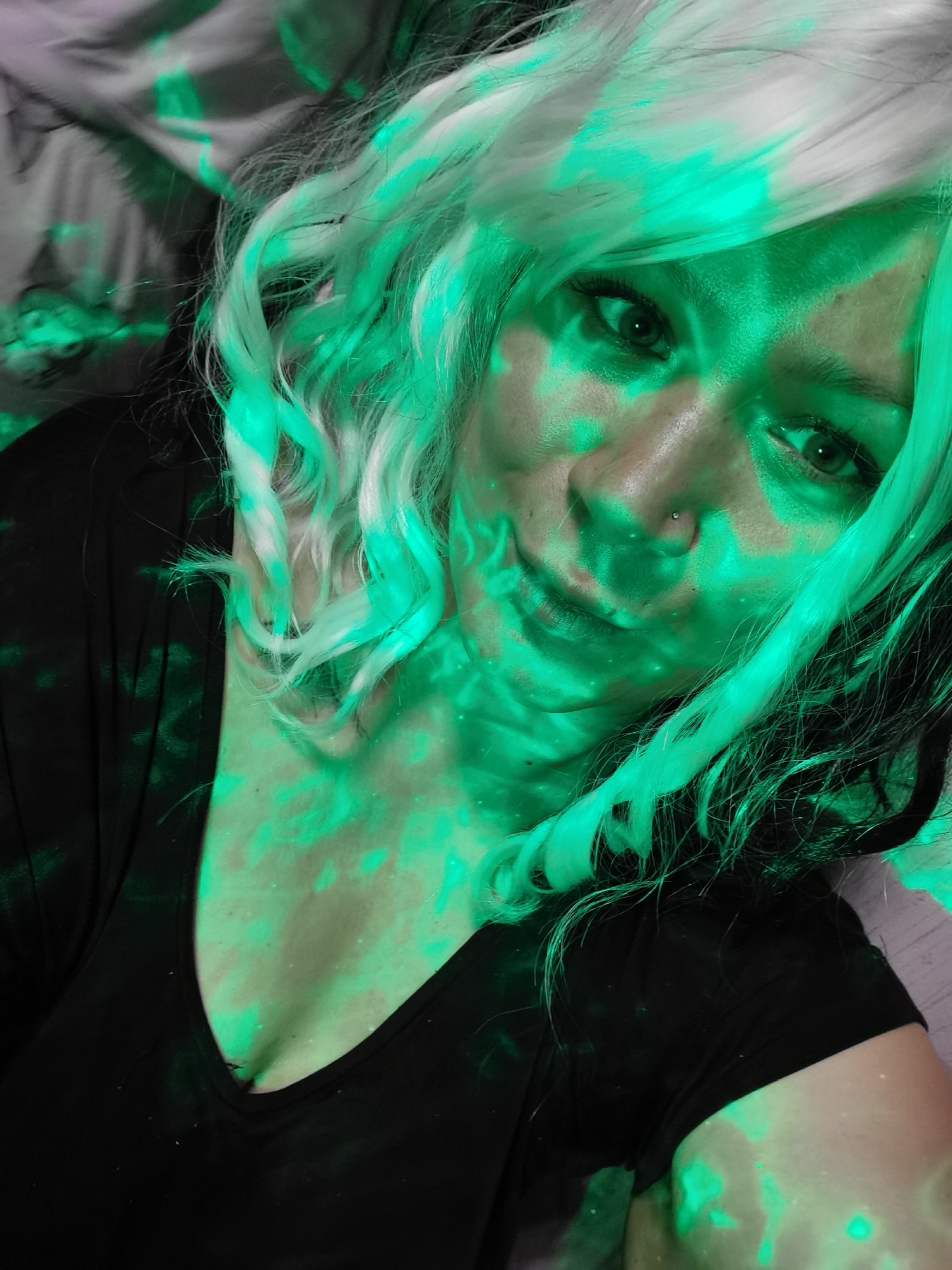 A medium close-up, canted, selfie-style shot shows a woman with fair skin and a nose ring. She wears a wavy wig that is mostly white with some darker strands mixed in. She is wearing a black v-neck shirt. She has dark eyes that are visible even with the green light. The background is mottled and indistinct, with both color and monochrome areas. The entire scene is bathed in an intense green light that obscures details and creates harsh shadows and highlights. I have no information available to me to determine if the image was captured in Votkinsk, Russia, or anywhere else for that matter.