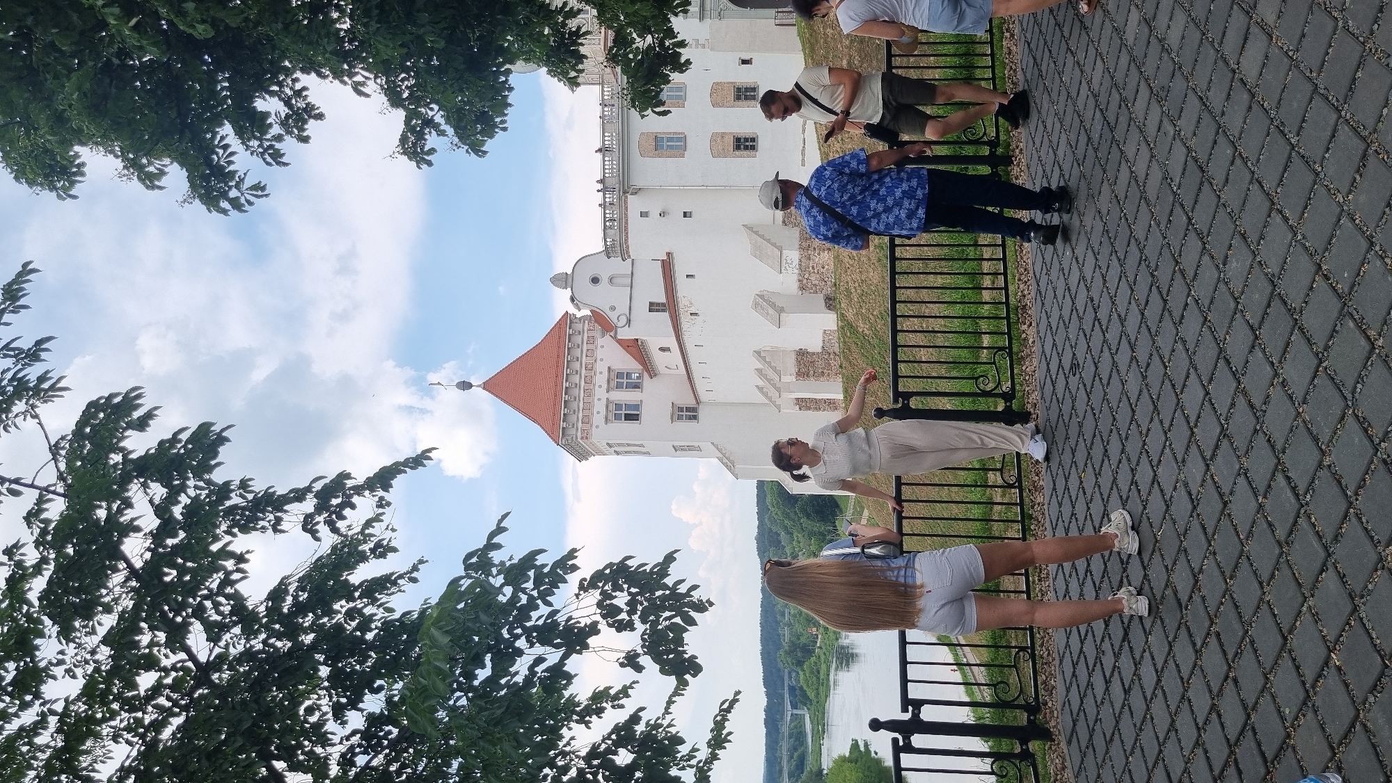 In Hrodna, Belarus, a group of people are gathered near the KAzimieradamm Palace. The palace, a large white building with a red-tiled roof and a prominent tower, is visible in the background. The foreground features a black metal railing, beyond which a river flows. A paved walkway with a diamond pattern is in the immediate foreground. Among the people, a woman with long blonde hair stands facing away from the camera, wearing a light blue top and white shorts. To her right, a woman in a white top and beige pants leans against the railing. Further to the right, a man in a blue patterned shirt and dark pants is visible, and next to him, a man in a light-colored t-shirt and khaki shorts holds a phone. Another person, partially visible on the far right, is wearing light blue shorts. The sky is partly cloudy.