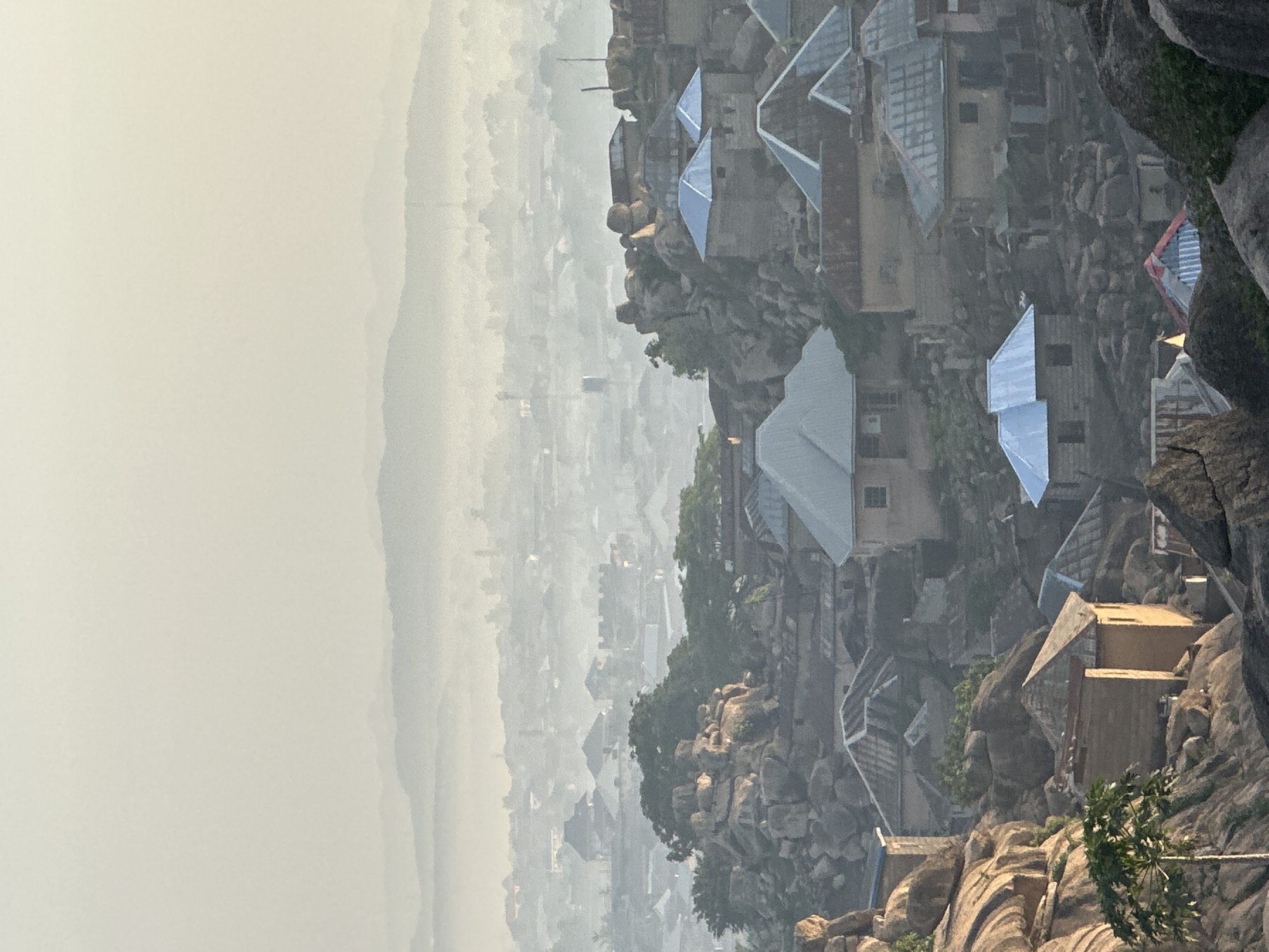 The image captures a unique urban landscape in Jos, Nigeria, characterized by settlements built directly into and around prominent rock formations. In the foreground and midground, numerous houses with varied rooflines, many appearing to be constructed with metallic or light-colored corrugated materials, are nestled among large, textured rock outcrops. The buildings exhibit a practical architectural style, adapting to the rugged terrain. Interspersed among the grey and brown rocks and structures are patches of green foliage, including trees and shrubs.
The background reveals a vast, sprawling city or town extending far into the distance. This urban expanse is largely obscured by a thick, atmospheric haze or fog, which gives the distant landscape a muted, almost monochromatic appearance, with faint silhouettes of buildings and hills barely visible against the pale sky. The sky itself is a uniform, light greyish-white, indicating either overcast conditions or a heavy atmospheric haze.
The diffused lighting suggests daylight hours, possibly early morning or late afternoon, given the hazy conditions. No people or specific activities are visible, presenting a static, panoramic view of this distinctive environment. There is no visible text in the image.