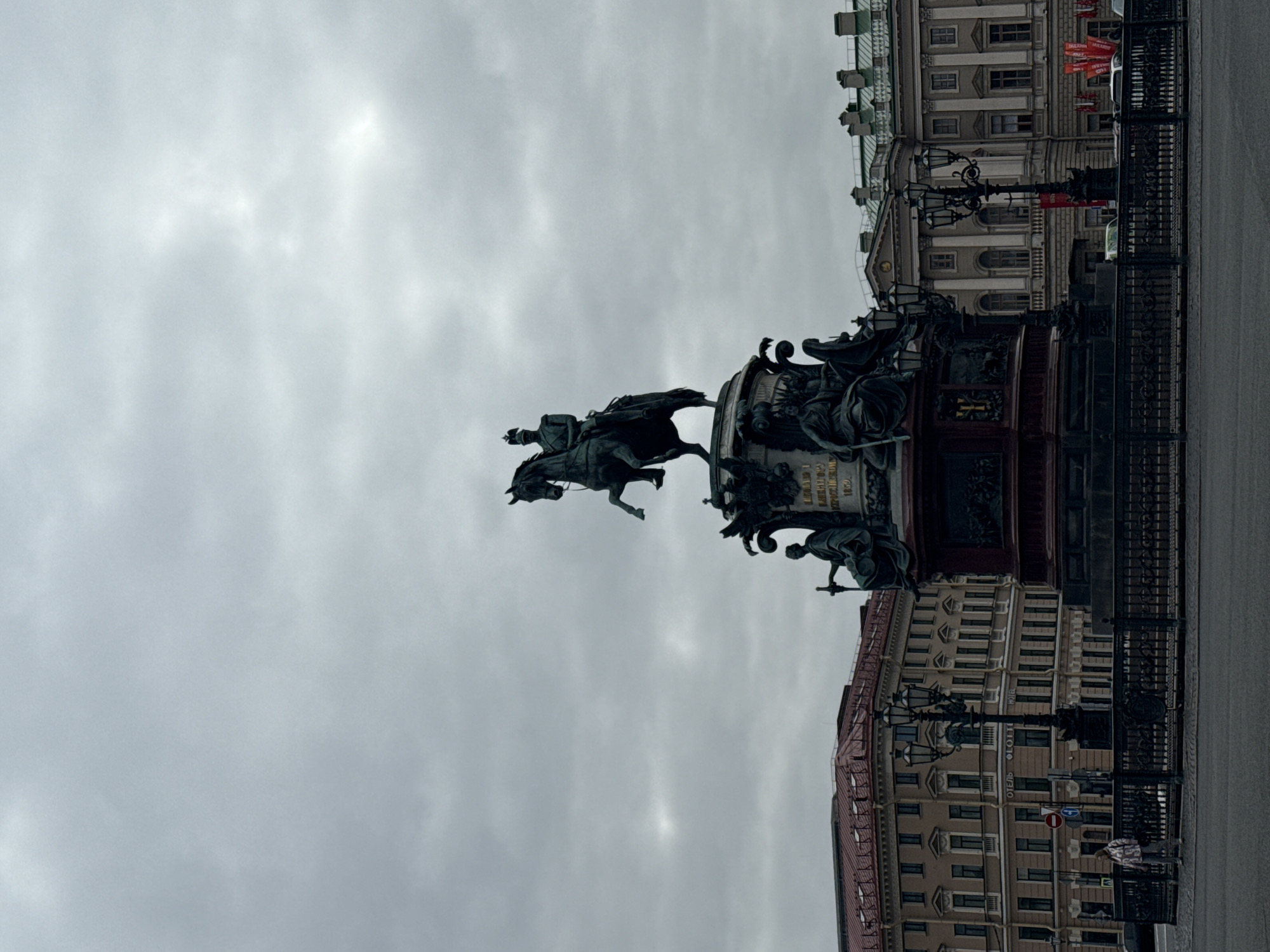 This image captures a grand equestrian statue dominating an overcast city scene in Saint Petersburg, Russia.
**Subjects**: The central subject is a large bronze equestrian statue of a male figure, likely an emperor or military leader, depicted on a rearing horse. The rider, dressed in military attire, extends his right arm forward. The statue rests on a tall, ornate pedestal adorned with relief sculptures and smaller, accompanying figures. Surrounding the monument are multi-storied, classical-style buildings with numerous windows and architectural details. One prominent building on the right (when viewing the image in its correct portrait orientation) clearly displays the word "ОТЕЛЬ" (Hotel). In the foreground, a dark, intricate metal fence and ornate street lamps are visible. A few indistinct human figures, likely pedestrians, can be seen in the distance near the buildings.
**Activity or Event**: The image depicts a static urban landscape, focusing on the architectural and sculptural elements of a city square or large thoroughfare. No specific activity or event is taking place beyond the general atmosphere of an urban environment.
**Notable Details**: The scene is illuminated by diffused light under a heavily overcast, cloudy sky, suggesting a muted daytime. The monumental scale of the statue and the classical architecture are characteristic of Saint Petersburg. The equestrian statue is identifiable as the Monument to Nicholas I on Saint Isaac's Square, a key landmark in the city.
**Visible Text**: The word "ОТЕЛЬ" (Hotel) is clearly legible on the facade of one of the buildings. On the pedestal of the statue, the dates "1825-1855" are visible, corresponding to the reign of Emperor Nicholas I.