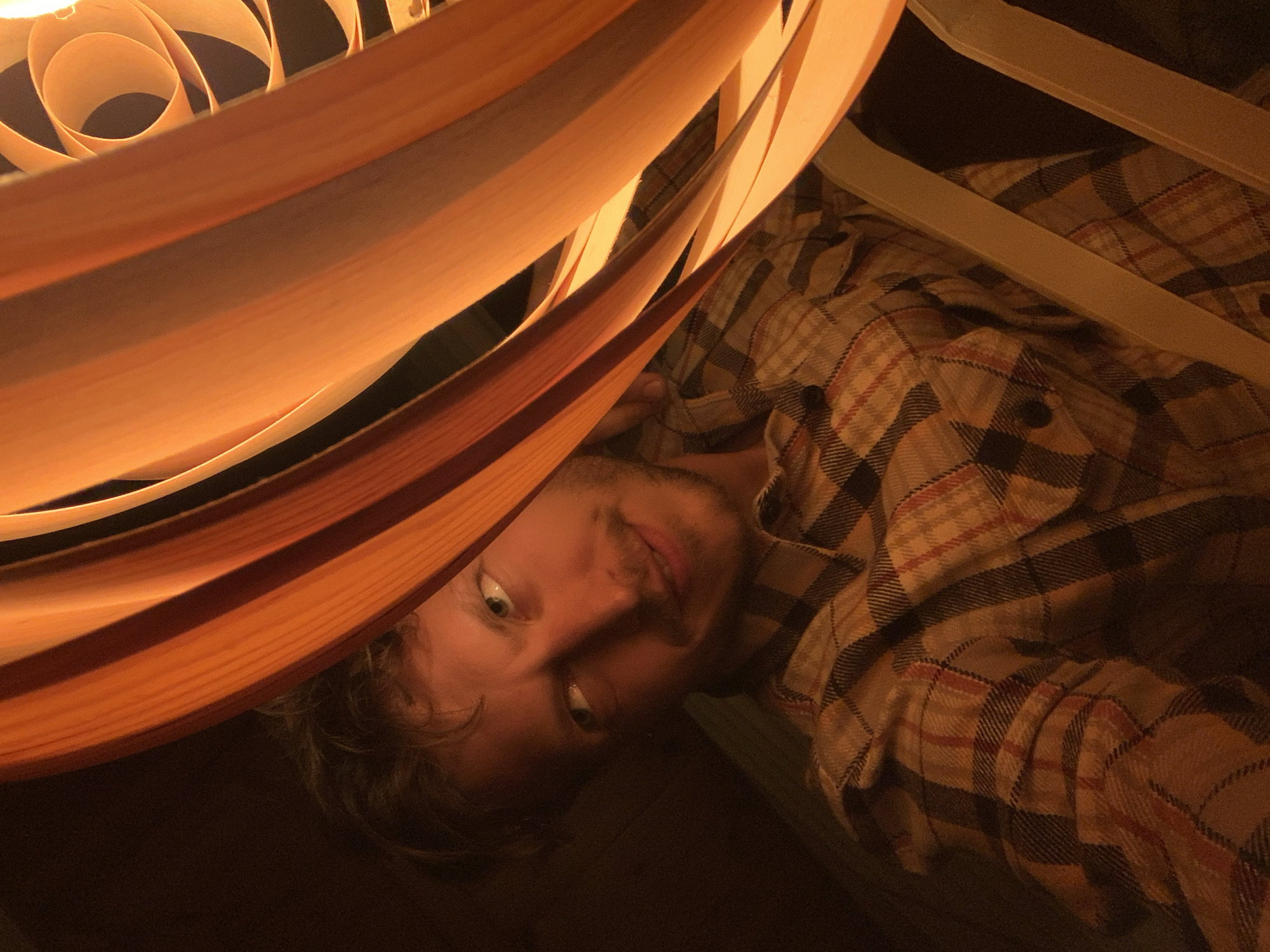 The image captures a close-up, low-angle view of a man lying down indoors, bathed in warm, artificial light. The man is positioned with his head tilted, looking directly at the viewer with wide, light-colored eyes and a neutral to slightly curious expression. He has short hair and some facial hair, possibly a mustache or stubble. He is wearing a plaid shirt with a pattern of beige, brown, and dark red or black lines.
A prominent feature in the foreground is a unique light fixture, likely a lamp shade, composed of overlapping, curved strips of a light-colored material, possibly wood or thick paper. This fixture emits a strong, warm orange-yellow glow, illuminating the scene and partially framing the man's face on the left side of the image. The strips create a layered, almost spiral effect.
Behind the man, a part of a bed or chair frame, appearing to be made of a light-colored material, is visible. The overall setting is dimly lit, suggesting evening or nighttime. The light source creates strong contrasts between the illuminated areas and the darker background. The image's orientation is tilted, adding to the dynamic perspective. No text is visible in the image. The provided location context is Tel-Aviv, Israel.