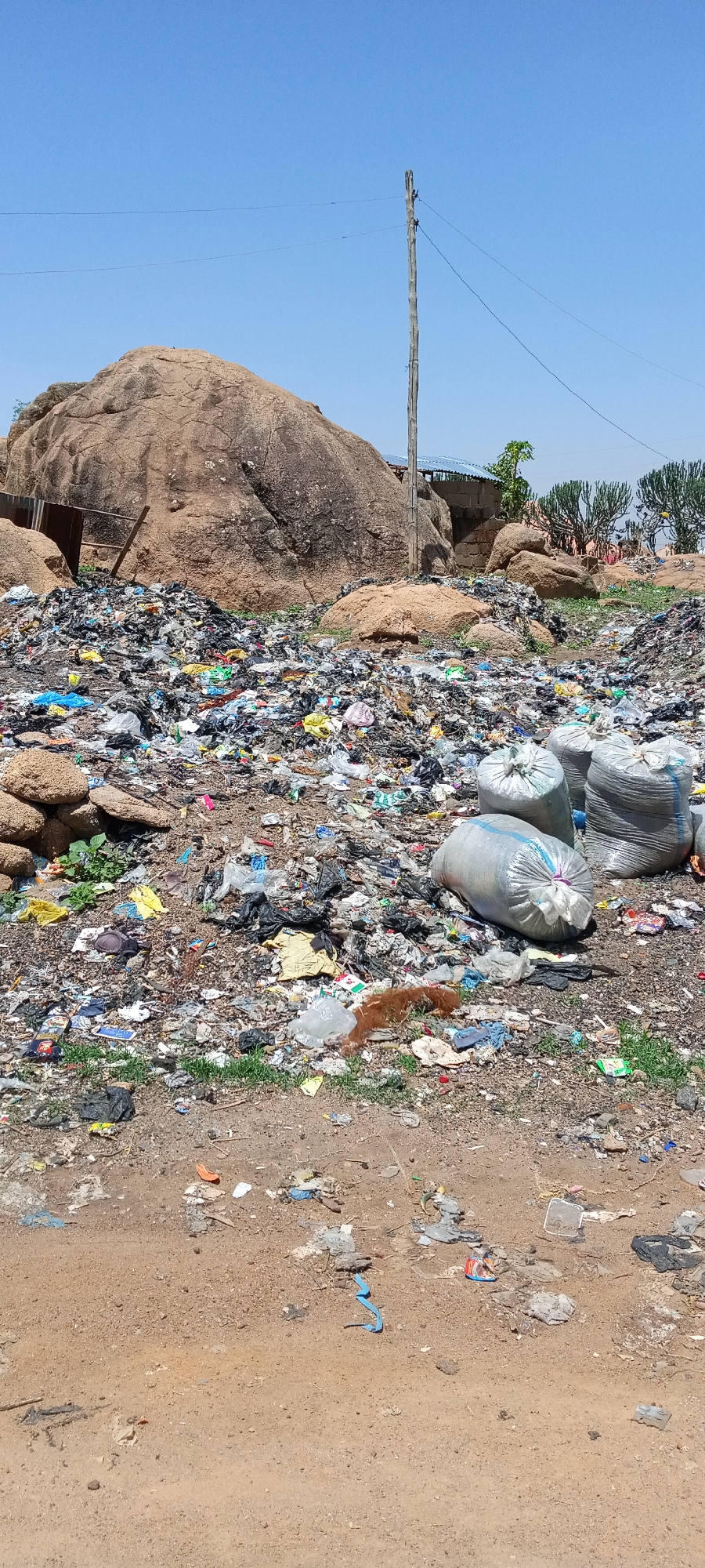 The image depicts a large, uncontrolled dumpsite in Jos, Nigeria. The foreground features a dirt road transitioning into a vast expanse of mixed waste, including plastic bags, bottles, fabric, and other discarded items. Three large, full bags of trash are prominently positioned on the right side of the frame. Behind the garbage, there is a massive rock formation, which is characteristic of the landscape in Jos. A utility pole stands in the background, along with glimpses of small buildings and vegetation. The sky is clear and blue, suggesting a sunny day. The overall impression is one of environmental degradation and improper waste management.