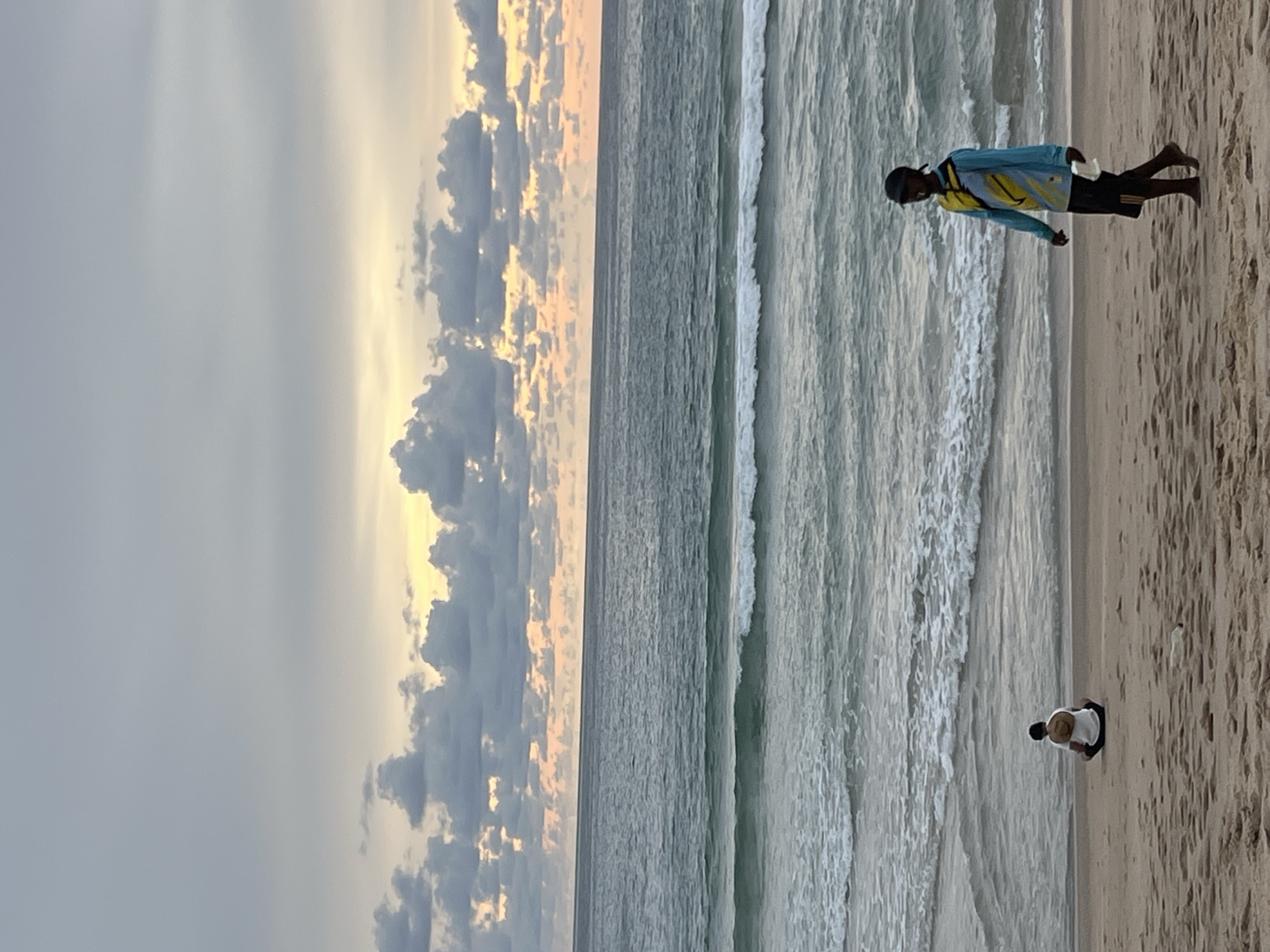 The image depicts a beach scene at what appears to be sunset or sunrise, given the soft, warm light illuminating the clouds. The sky is a mix of gray and soft yellow, with fluffy clouds present. The ocean is calm with gentle waves rolling onto the sandy shore.
There are two people visible on the beach. One individual is standing closer to the water, wearing a light blue long-sleeved shirt with yellow accents and dark shorts. They are facing away from the camera, looking towards the ocean. The second person is further up on the sand, appearing to be kneeling or sitting, wearing a white top and dark shorts, with a straw hat on their head.
The setting is a sandy beach with the ocean stretching out towards the horizon. The lighting suggests it is either early morning or late evening. There are no visible objects other than the people and natural elements of the beach. No text is visible in the image.