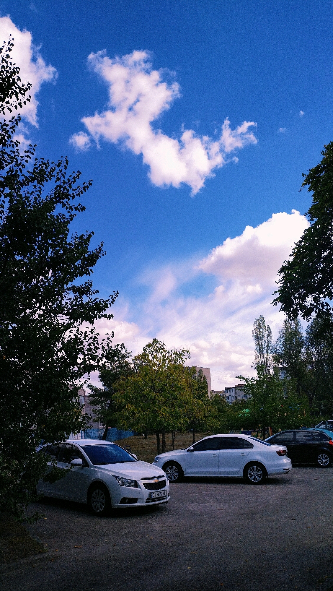 A low-angle, full shot captures a bright, partly cloudy sky above a residential area in Horishni Plavni, Ukraine. The sky is a vibrant blue, populated with scattered white clouds, some wispy and streaky, others more voluminous. The tops of dark green trees frame the left and right sides of the image, their branches reaching towards the sky. In the middle ground, more trees with lighter green foliage are visible, along with several buildings, some appearing to be apartment complexes. A paved parking area occupies the foreground, where three cars are parked. The car closest to the viewer is a white sedan. To its right is another white sedan, and further back, a dark-colored sedan is parked. The scene appears to be a typical urban landscape on a clear day.
