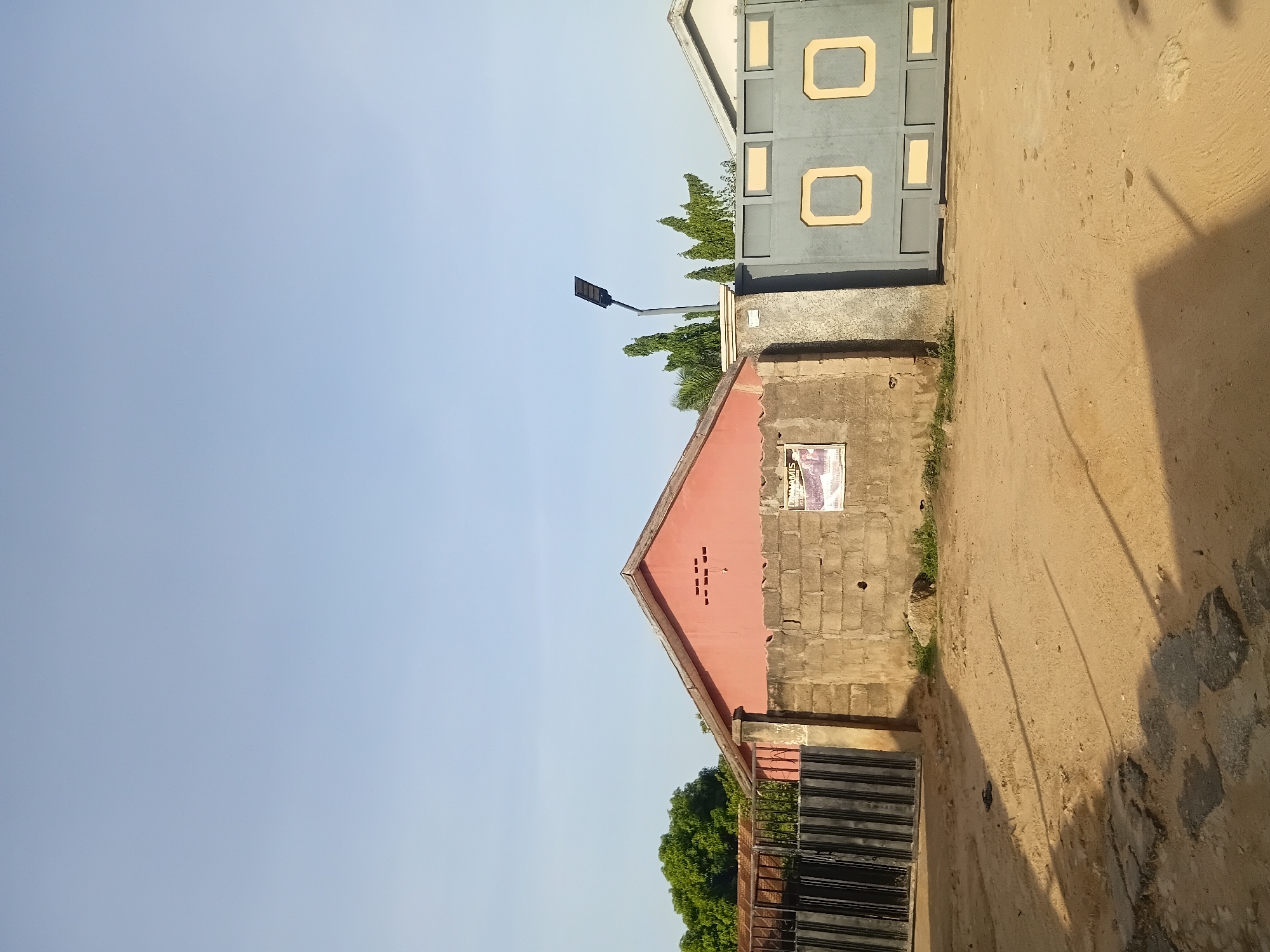 The image depicts the exterior of a property in Jalingo, Nigeria. The scene is dominated by a dirt road in the foreground, with patches of broken asphalt. Behind the road, there is a wall constructed from concrete blocks, with a red-painted building visible beyond it. The building has a gabled roof and a few small rectangular openings near the peak. A black metal gate is situated to the left of the wall, and a larger, gray metal gate with yellow square accents is to the right. A solar-powered street light stands on a pole to the right of the wall, near the gray gate. Lush green trees are visible behind the structures. The sky is clear and blue. A single sandal lies on the dirt road, casting a shadow. A poster is affixed to the wall.