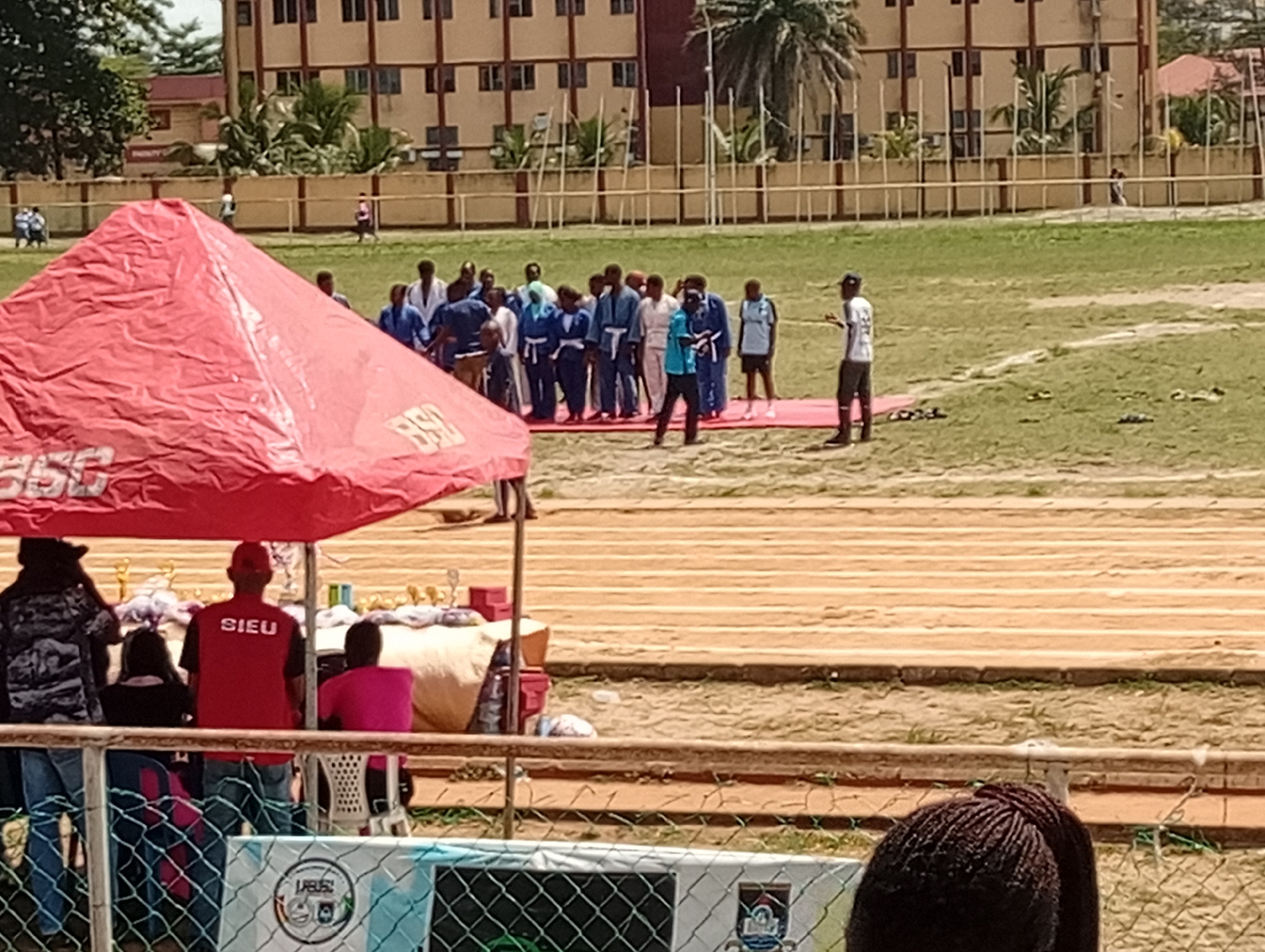 A sunny outdoor scene features a group of people on a grassy field in front of a tan-colored building in an unknown city. A group of people dressed in athletic wear, predominantly blue and white, are standing in a loose line, possibly preparing for an event or having just completed one. Two men dressed in dark clothing appear to be giving instructions or speaking to the group.
In the foreground, a red tent structure partially obscures the view of a table holding trophies and various items. A person wearing a red shirt with the letters "SIEU" on the back is standing near the table. A chain-link fence is visible in the lower portion of the frame, and behind it, a partial view of a person's hair. The field shows visible track lines marked in light brown or tan.