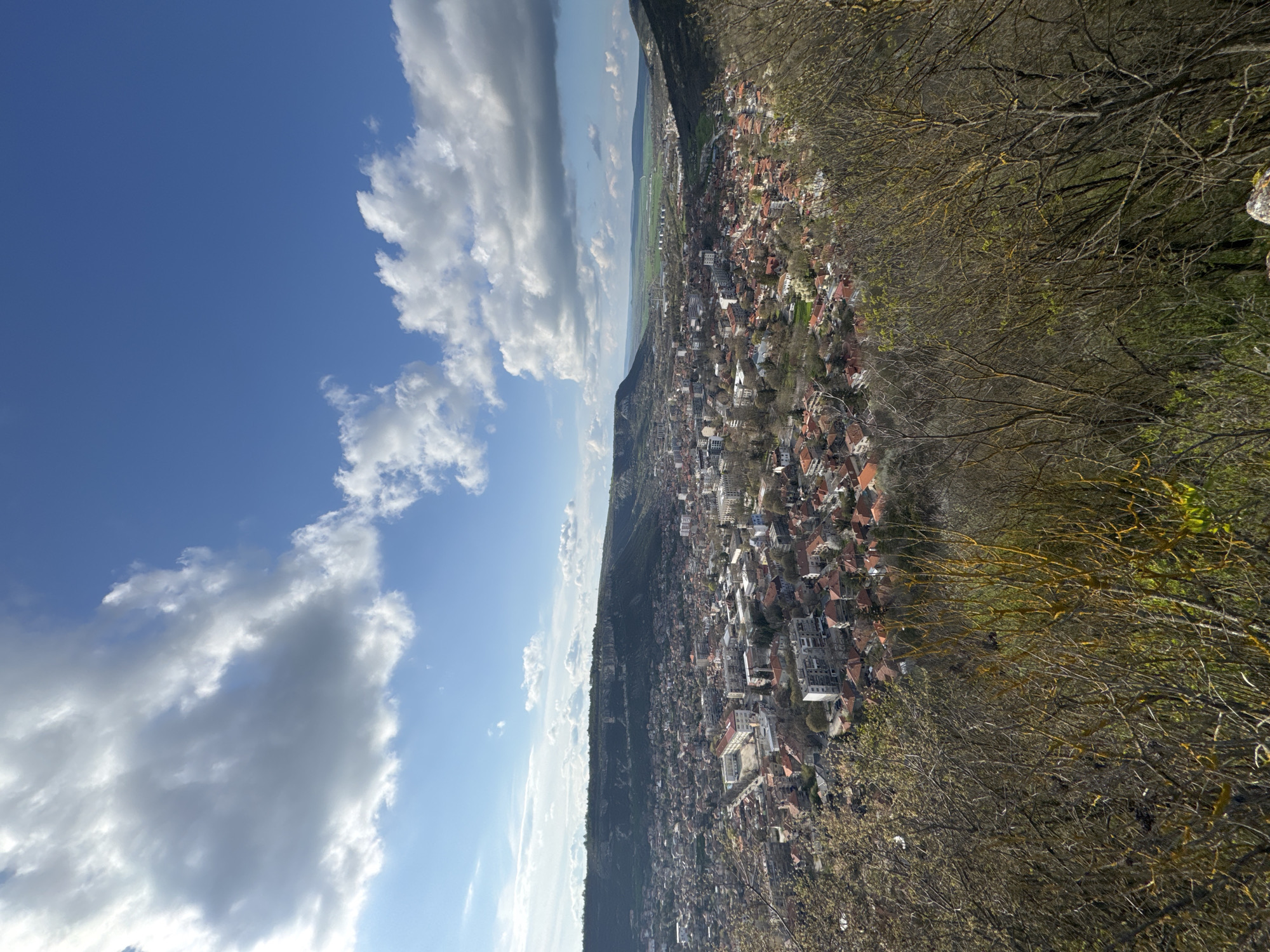 The image presents a landscape view of Provadia, Bulgaria, taken from an elevated perspective. The sky occupies the left portion of the image and is filled with a blue expanse adorned with cumulus clouds. A ridge line runs diagonally across the center of the image, separating the sky from the urban sprawl. The urban area consists of buildings with predominantly red-tiled roofs, interspersed with greenery. The composition is framed on the right by trees, their branches extending into the scene. The overall scene suggests a spring or early summer day with clear weather conditions. The city is nestled at the foot of a ridge, which extends towards a green area, and is lined with buildings stretching along the terrain.