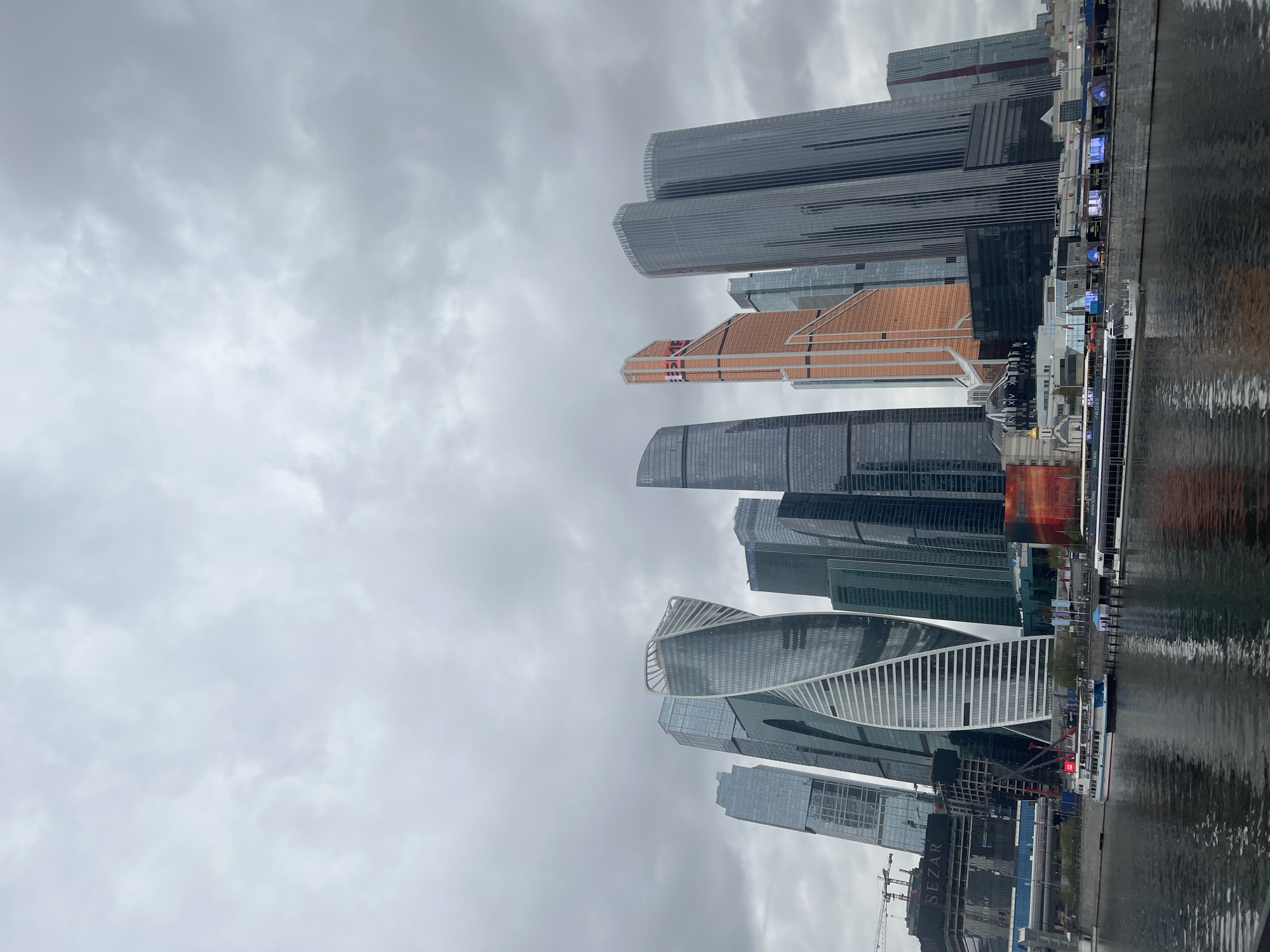 This is a vertically oriented photograph of the Moscow International Business Center (also known as Moscow City) reflecting in the Moscow River, Russia. The overcast sky above the cityscape consists of dense, grey clouds. In the foreground, the water reflects the city's skyline. Several distinct skyscrapers are visible, including the Evolution Tower (with its twisted design), the Mercury City Tower (with its copper-colored exterior), and other prominent structures with varying heights and architectural styles. Along the riverbank, a construction site is visible with a crane and partially built structures. A river cruise boat is docked along the embankment.