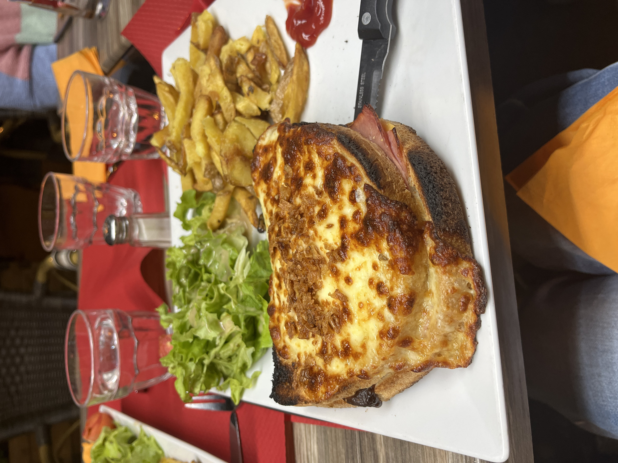 A close-up, medium-angle shot displays a meal on a white, rectangular plate. The meal consists of a croque monsieur, golden-brown potato wedges, a small portion of green salad, and a dollop of ketchup. A black-handled knife rests on the plate next to the sandwich. Three glass tumblers are positioned to the left of the plate on a red placemat. The meal appears to be served at a restaurant in Montélimar, France, based on common dishes in the region. A person wearing denim jeans and an orange garment is visible on the right side of the image.
