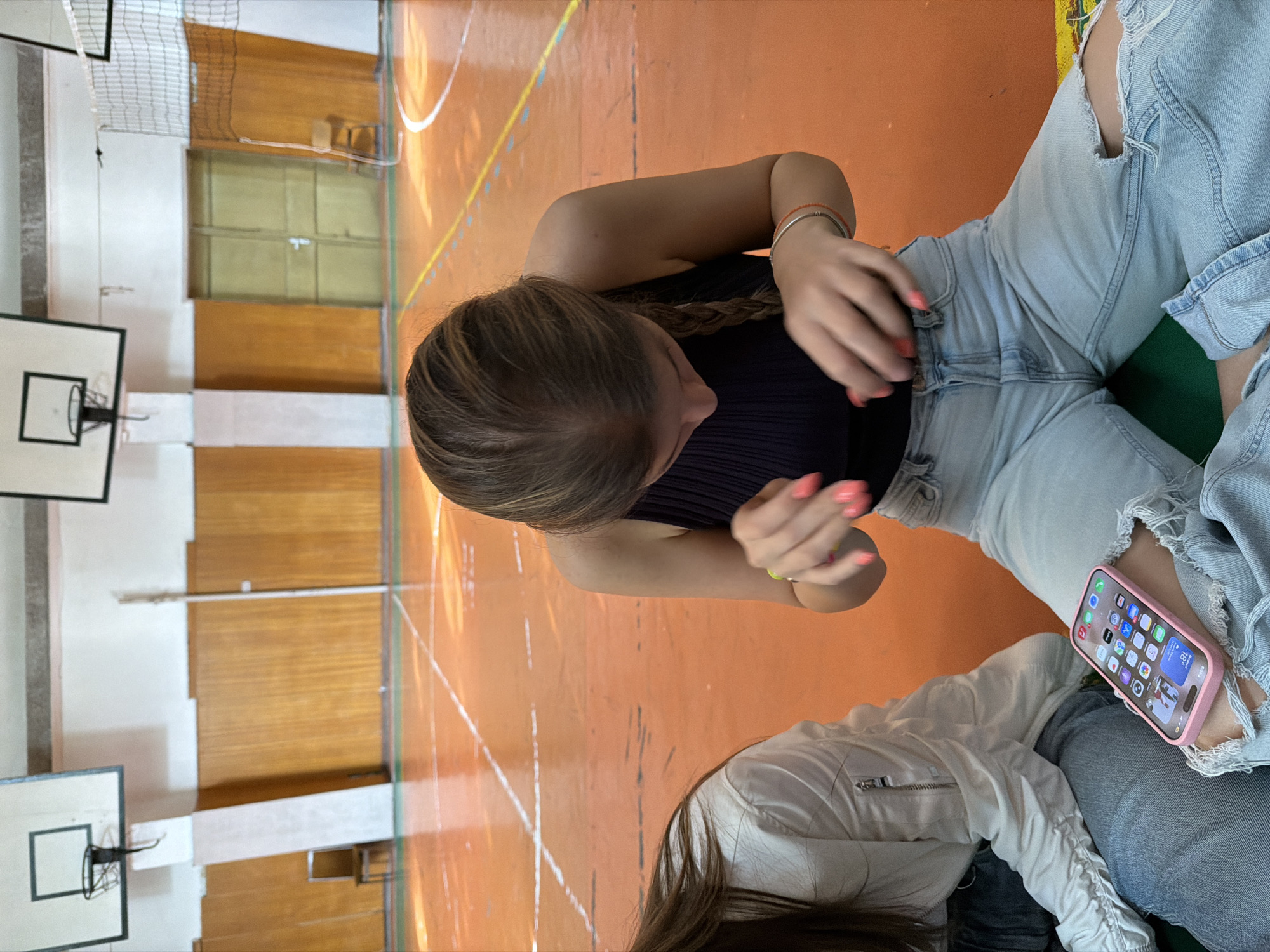 The image depicts two individuals resting in an indoor sports hall in Burgas, Bulgaria. The photograph is taken from a high angle, looking down upon them.
In the foreground and center, a young woman is prominently featured, seen from the top of her head down to her waist. She has light brown hair styled in a braid. She is wearing a black ribbed sleeveless top and light-wash ripped jeans. Her hands, adorned with bright pink nail polish, are clasped in front of her, and she wears a red beaded bracelet and a silver bracelet on her left wrist. She is looking downwards.
Partially visible in the bottom right corner is another individual, seen from the back of her head and shoulders. She has dark hair and is wearing a white jacket with a zippered pocket, paired with light-wash jeans. She is holding a pink-cased iPhone, which displays various app icons, the time "18:20", the day "Wed 4", and a temperature of "18°" on its screen.
The setting is a sports gymnasium with a reddish-orange wooden floor marked with white and yellow lines for different courts, including basketball and possibly volleyball or badminton. Two basketball backboards with hoops are visible against a white wall with wooden paneling on the left side of the frame. A net, likely for volleyball or badminton, is partially visible in the background. The overall activity appears to be a casual break or observation rather than active participation in sports. The lighting suggests daytime indoors.
