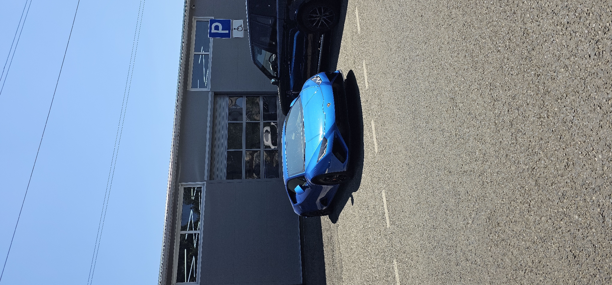 An eye-level, slightly angled, full shot captures a bright blue Lamborghini Huracan parked on an asphalt surface. The sports car is positioned in the foreground, facing slightly to the right. Behind it, to the right, is the rear portion of a dark-colored SUV. The backdrop is a gray building with a large, segmented glass garage door and a smaller window to its left. A blue and white parking sign, indicating a disabled parking space, is mounted on the building to the right of the garage door. The sky is clear and blue, with power lines visible across the top of the frame. The lighting suggests a bright, sunny day. This scene is located in Krasnodar, Russia.