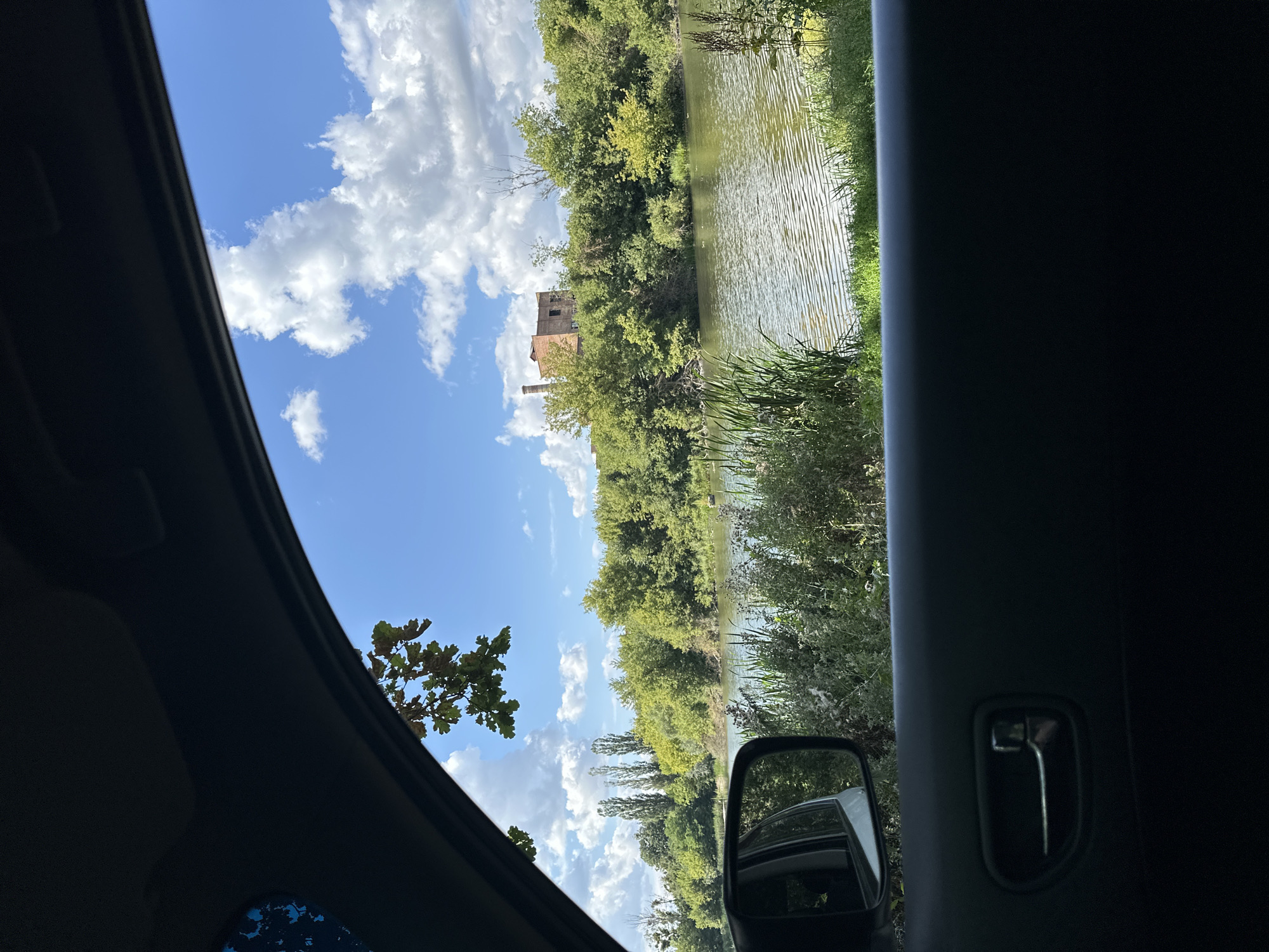 The image, taken from inside a vehicle, looks out at a calm river bordered by lush green trees and vegetation. A weathered, brick building with a prominent chimney stands on a rise behind the trees. The sky is a vibrant blue, dotted with fluffy white clouds. A side-view mirror of the car is visible on the left, reflecting the scene. The foreground shows the interior of the car, including a door panel and a door handle. The location is the city of Shatava, Ukraine.