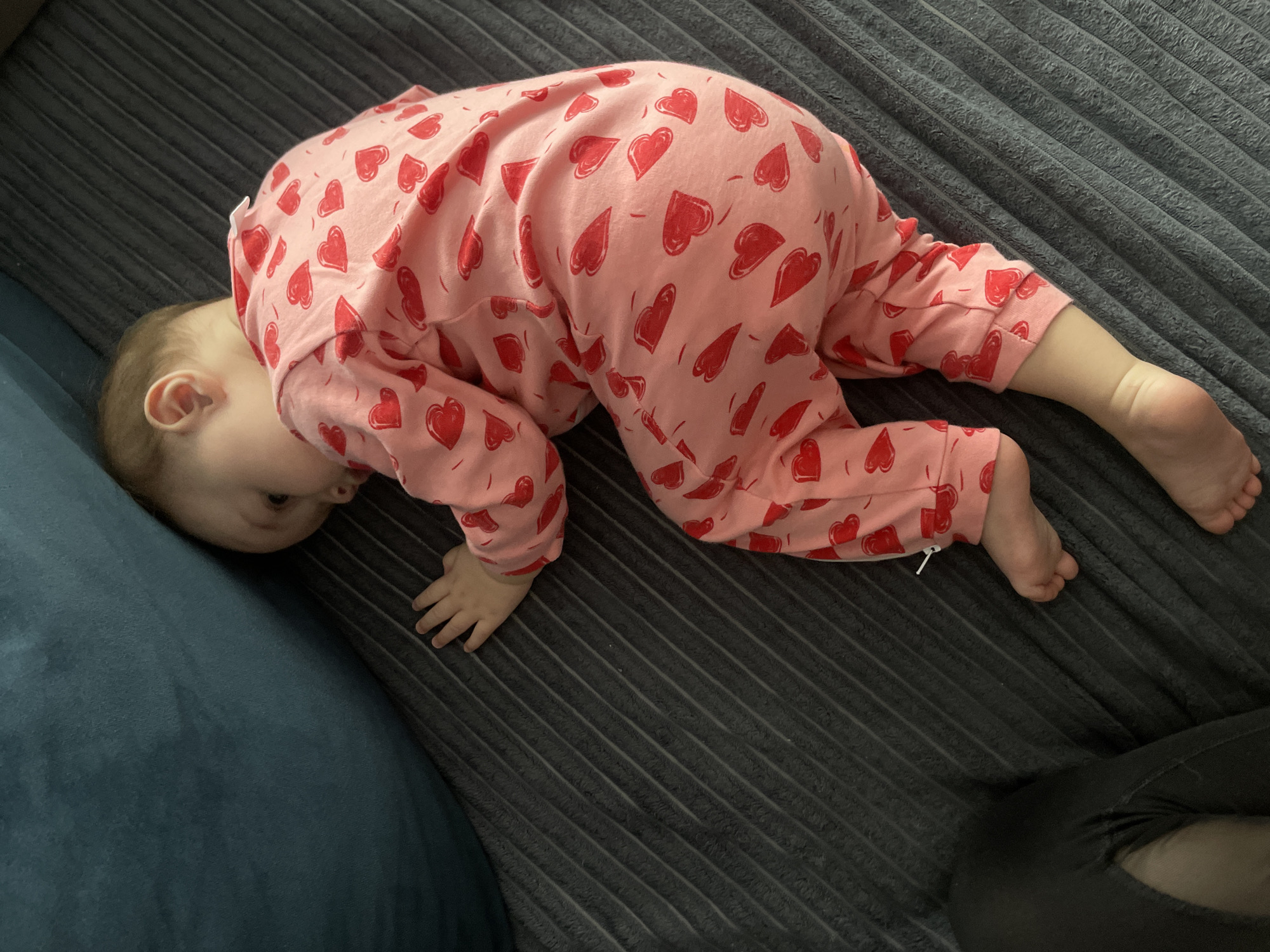 An eye-level, indoor shot depicts a baby or young toddler positioned on a dark gray ribbed surface, likely a sofa or blanket. The child is wearing light pink pajamas adorned with numerous red heart patterns. The baby is in a bent-over or crawling stance, with its head tilted down and resting on a dark blue cushion or a different section of the sofa, which occupies the left side of the frame. Its hands are placed on the gray ribbed surface for support, and its bare feet are visible towards the right. One of the baby's eyes is partially visible, suggesting a focused or curious expression as it looks downwards. In the bottom right corner, a glimpse of dark clothing, possibly an adult's leg or pant, can be seen. The lighting in the image is soft and ambient, indicative of an indoor daytime setting. The scene captures a moment of the child exploring its surroundings or engaging in play. The location context for this media is Подшивалово, Russia. No visible text is present in the image.