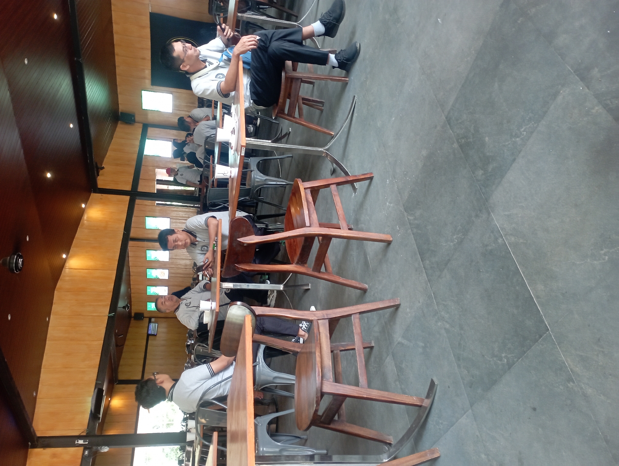 In the city of Bajulan, Indonesia, a group of men are seated at tables in what appears to be a cafe or restaurant with a rustic interior design featuring exposed wooden beams and paneling on the ceiling and walls. The flooring is composed of large, dark grey stone tiles.
Four men are prominently visible, dressed in light-colored collared shirts and dark trousers, suggesting they might be colleagues or attending an event. One man on the far right is looking at his phone, while another man in the center-right is also looking at his phone. Two men are seated to the left, facing away from the camera, with one looking towards the other.
The tables are set with what appear to be cups, plates, and potentially food or drinks. Several chairs are visible, including wooden chairs with a classic design and more modern metal chairs. The overall atmosphere suggests a casual gathering or break in a semi-formal setting. The lighting in the room is a mix of natural light from large windows and artificial lights from the ceiling.