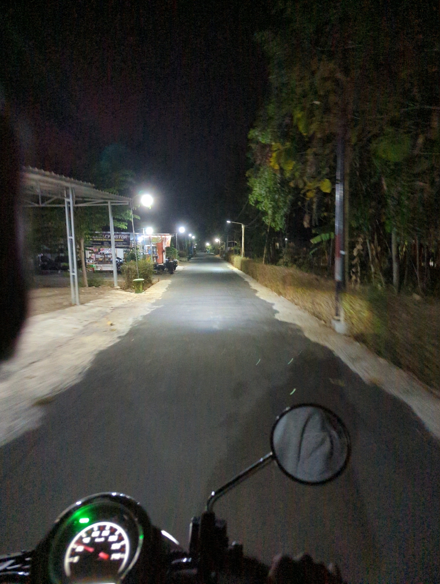 The image captures a first-person perspective of someone riding a motorcycle at night on a paved road in Kedungdowo, Indonesia. The road is illuminated by streetlights, casting pools of light and long shadows. On the left side of the road, there is a covered area with support poles, possibly a small shop or stall, with signs visible. Beyond this structure, trees and foliage are dimly lit. On the right side of the road, dense vegetation lines the path, with a single streetlight in the distance. The motorcycle's dashboard, including a speedometer with a green indicator light, is visible in the foreground. A side mirror reflects a gray object, possibly fabric. The overall atmosphere is dark, with the artificial lighting highlighting portions of the environment.
