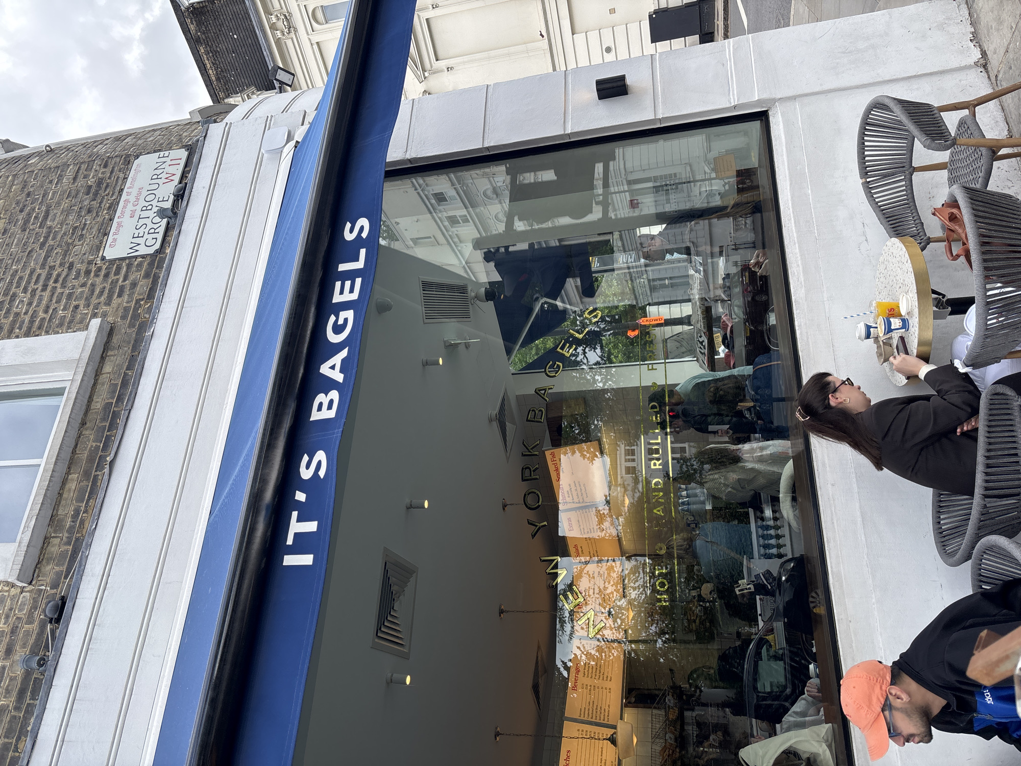 The image shows the storefront of "It's Bagels," a New York-style bagel shop. The shop is located on Westbourne Grove, W.11, in the Royal Borough of Kensington and Chelsea in London, United Kingdom. The awning of the shop is blue, and the name of the shop is written in white letters. The shop has a large glass window that reflects the surrounding buildings and people inside. Outside, there are tables and chairs where people are seated, including a woman in a black blazer and a man with an orange cap. The menu is visible on the window with the words "New York Bagels" and "Hot Hand Rolled Fresh."
