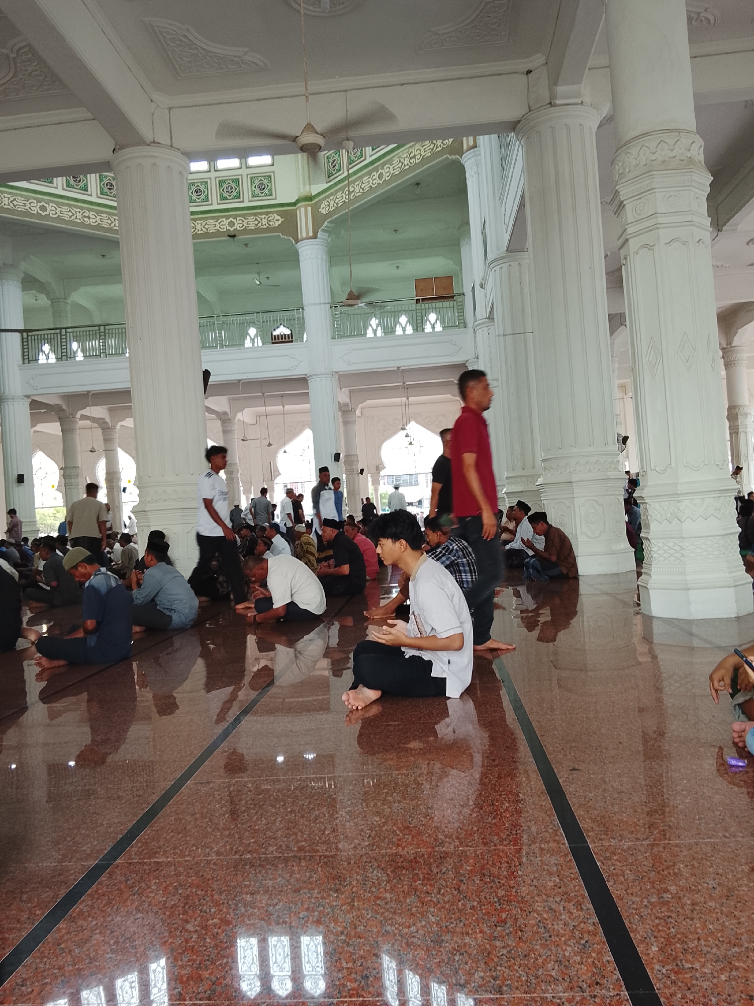The image shows a group of men sitting on the polished floor of a mosque in Lhokseumawe, Indonesia. The mosque has high ceilings with decorative molding and is supported by large white pillars. Fans are visible on the ceiling. The floor is made of red granite with dark lines. Some men are seen praying or in quiet contemplation, while others appear to be conversing. The overall atmosphere suggests a place of worship and community gathering.