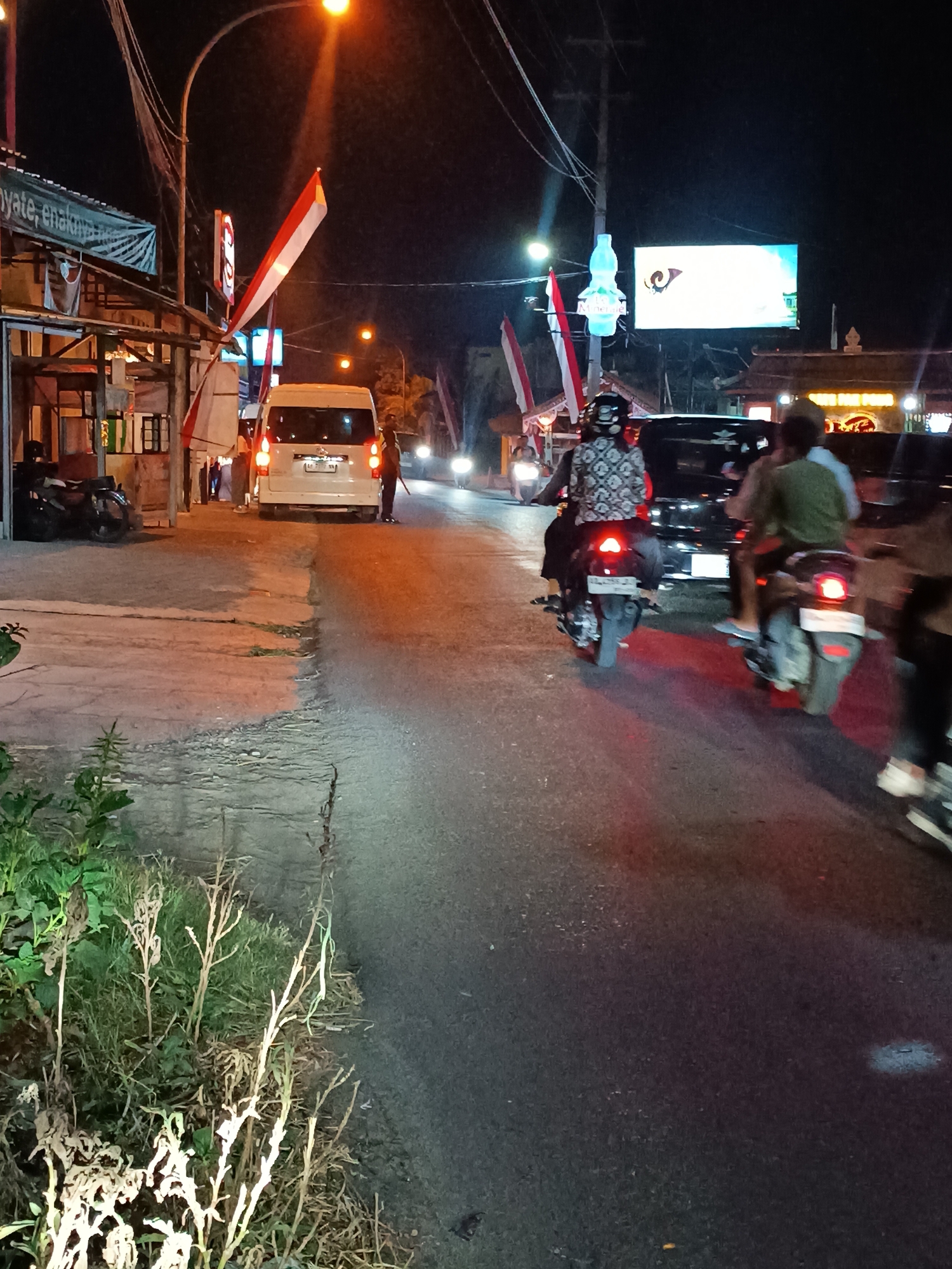 An eye-level, night time, medium shot captures a street scene in Pleret, Indonesia. The paved road occupies the right two-thirds of the frame, while the left third shows a raised concrete sidewalk with some vegetation. A white van is parked on the left side of the road, facing away from the viewer, with its brake lights illuminated. Two Indonesian flags are prominently displayed, one on a pole near the van, and another larger one attached to a building on the left. Several people are on motorcycles, moving along the road. On the left side of the road, a person stands next to the van. Two individuals are riding a motorcycle in the foreground, facing away from the viewer, their brake lights are on. Another person is riding a motorcycle slightly behind them, also facing away. A large, illuminated billboard is visible in the background. The scene is illuminated by streetlights and the lights from vehicles and the billboard.